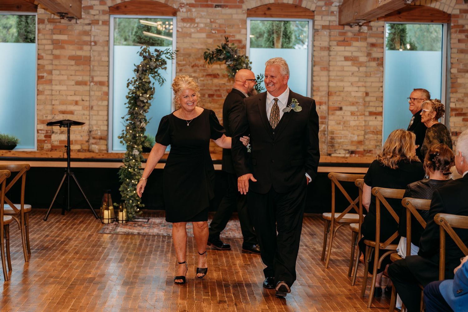 Elegant couple walking down wedding aisle in brick venue, man in suit with boutonniere, woman in black dress.