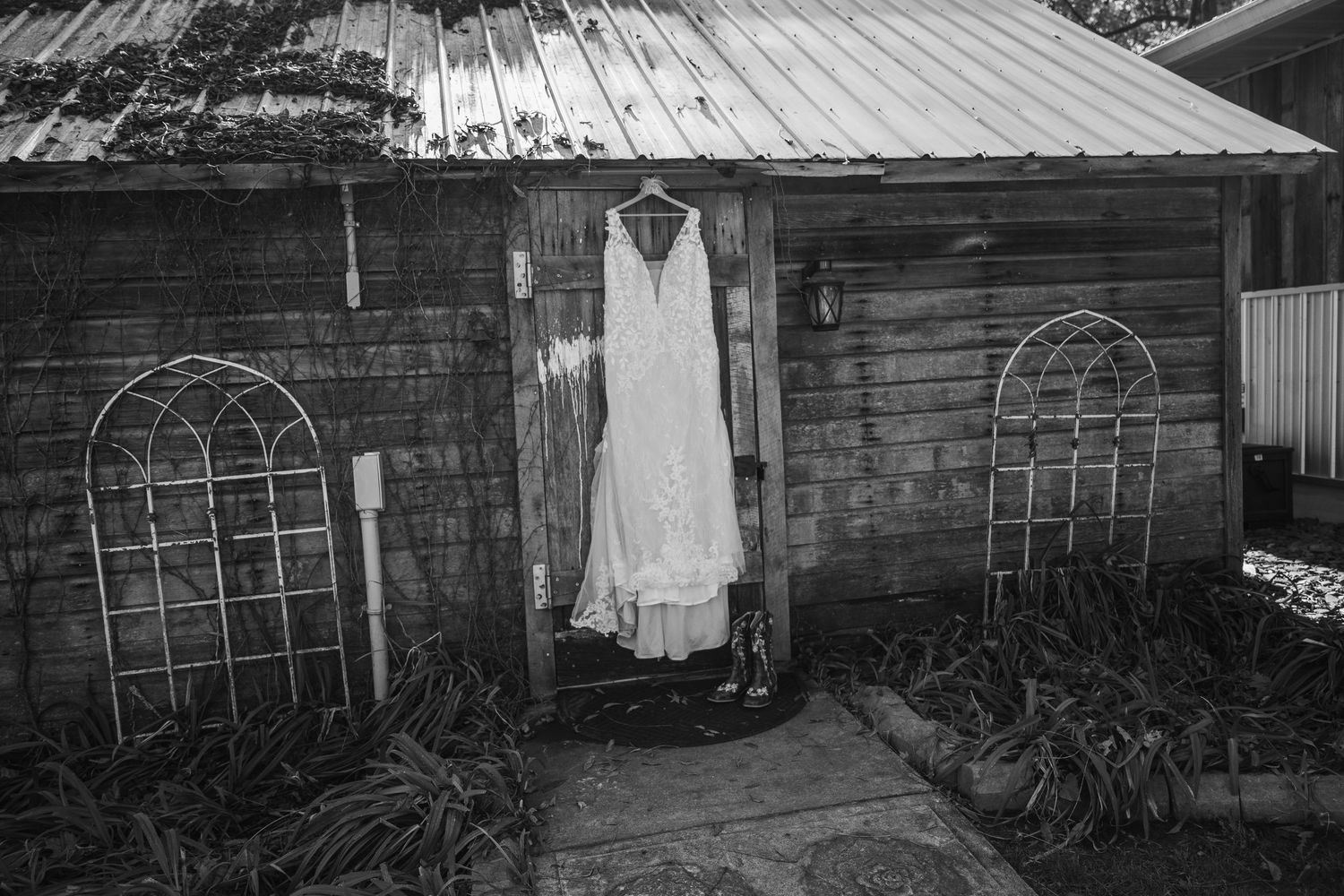 Black and white photo of a wedding dress hanging on a rustic wooden shed door, surrounded by garden decor.
