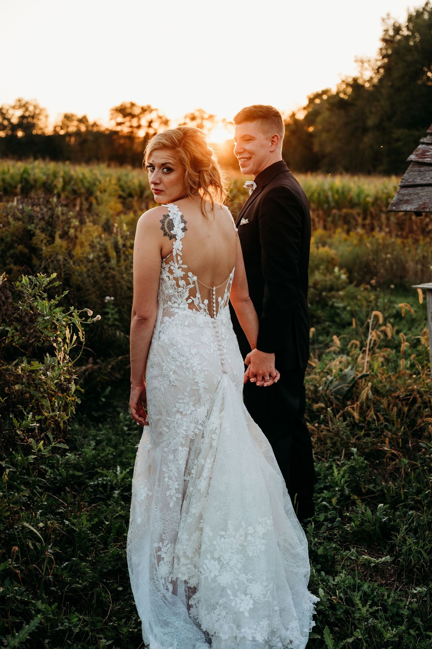 A couple in wedding attire hold hands at sunset in a wildflower field, with the bride wearing a lace backless gown.