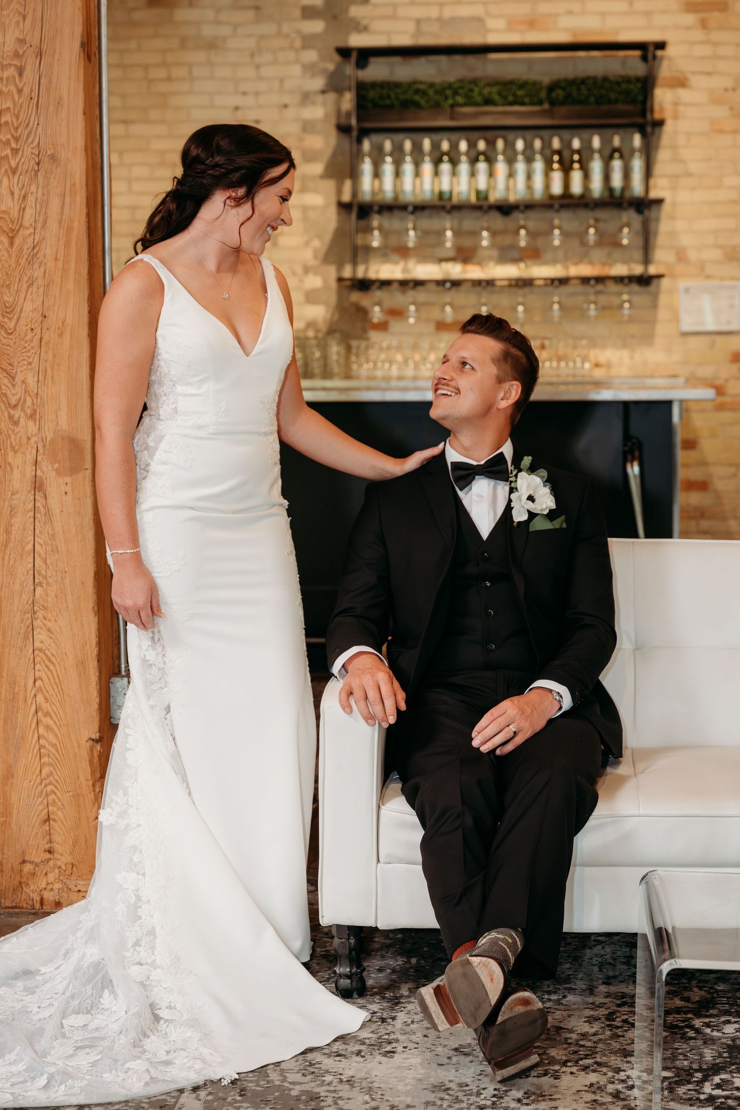 A couple in wedding attire share an intimate moment while seated on a white couch in an industrial-style space.