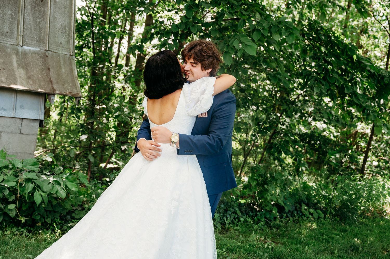 A bride in a white backless gown embraces a groom in a navy suit outdoors surrounded by lush green trees.