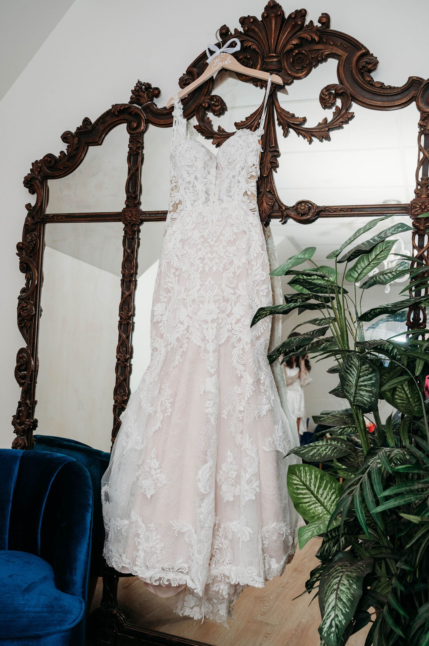 A white lace wedding dress hangs from an ornate brown wooden mirror frame with green plants in the background.