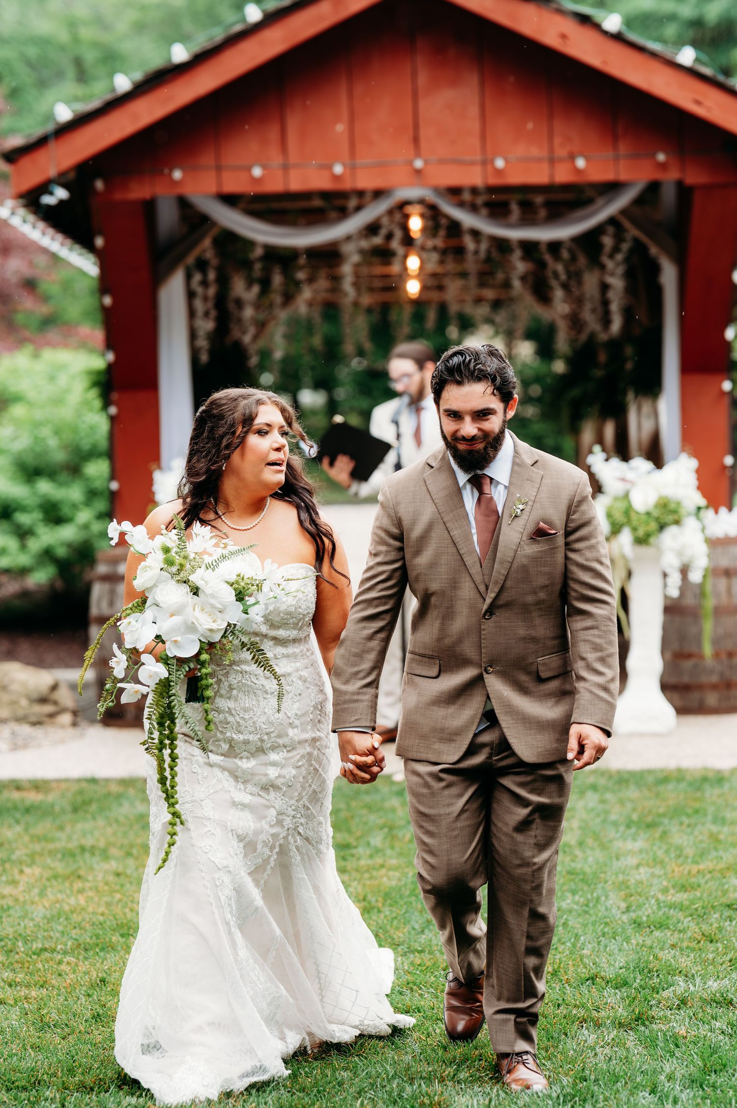 Bride and groom walk hand-in-hand on green lawn before a rustic red barn decorated with string lights.