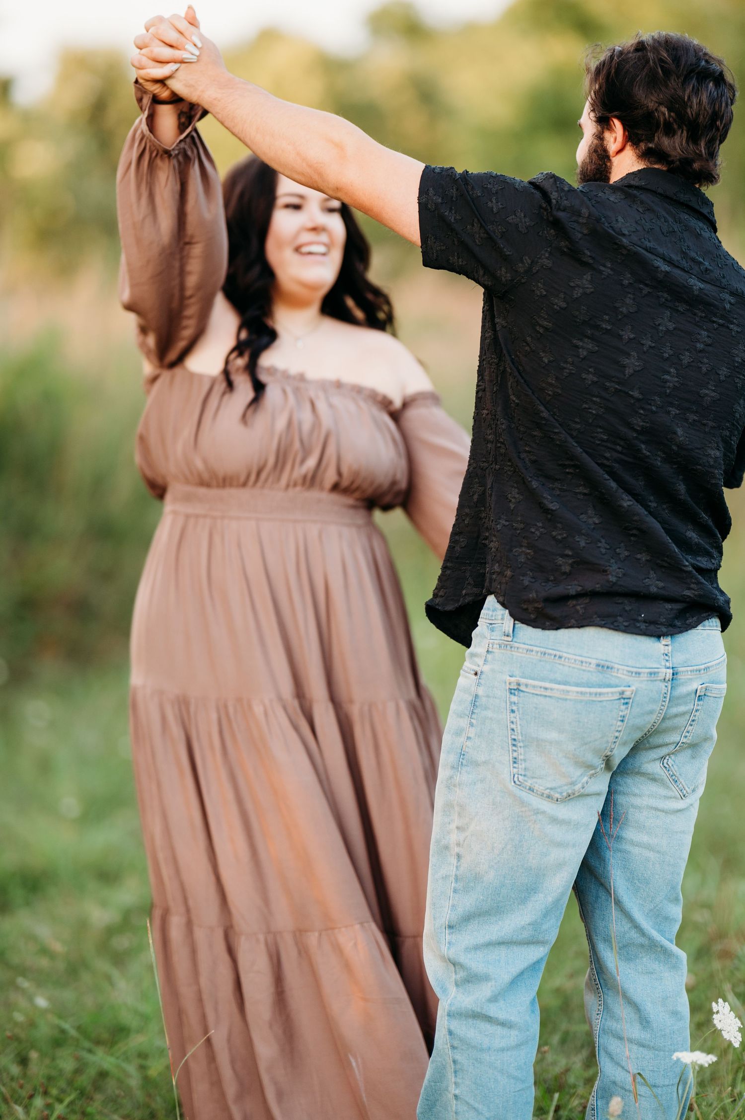 A couple dances outdoors in a green field, woman in flowing brown dress smiling as man in black shirt twirls her.