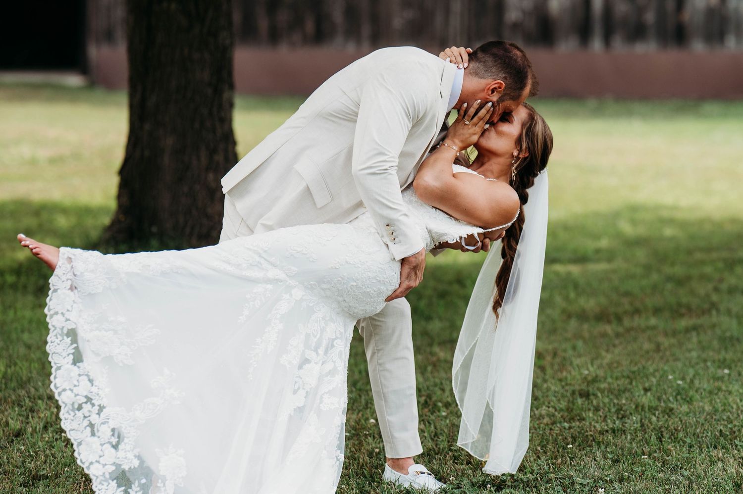 Groom dips bride for a romantic kiss outdoors on green grass, bride wearing white lace gown and veil.