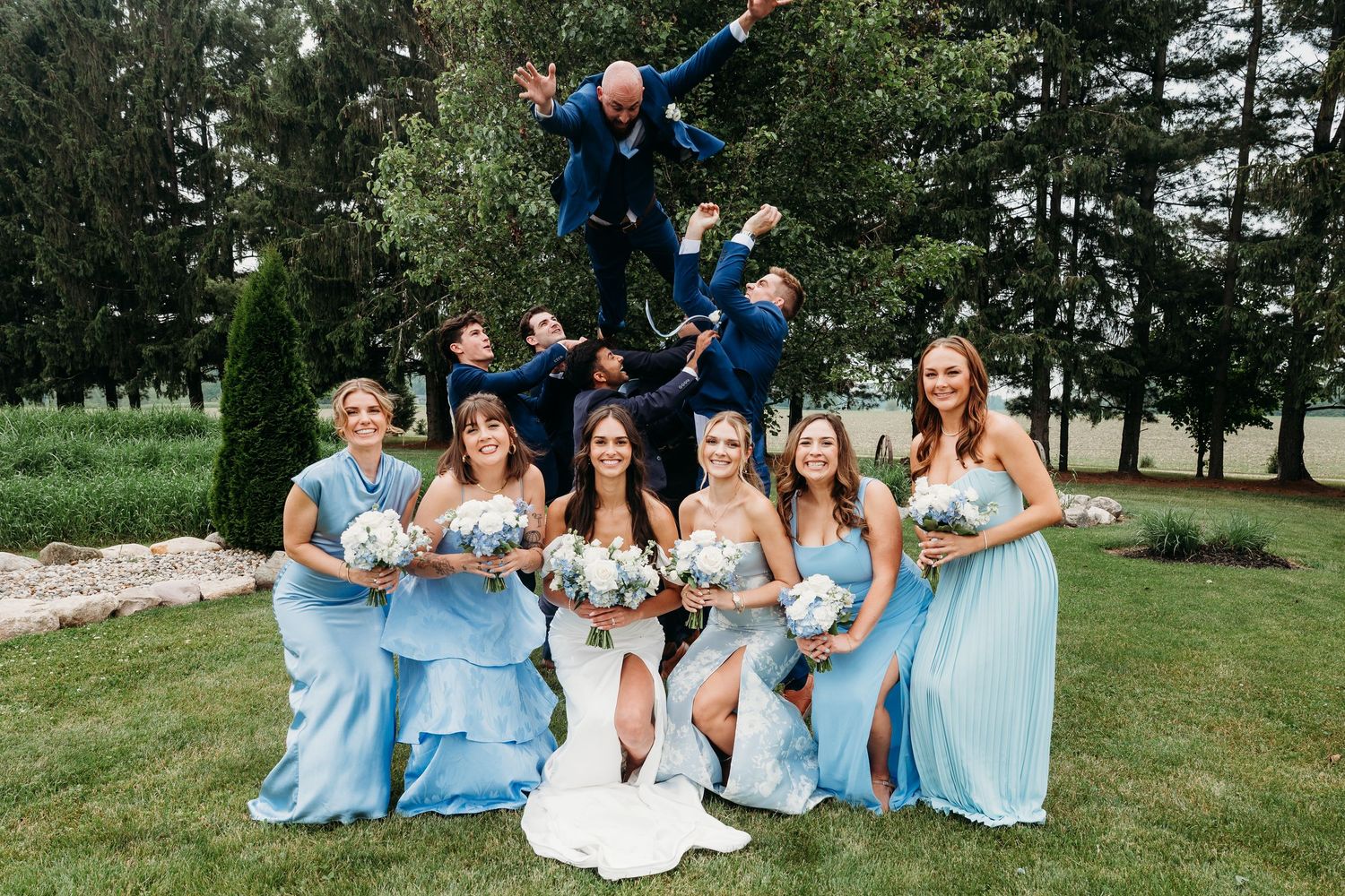 Bride and bridesmaids in light blue gowns holding bouquets while groomsmen leap joyfully behind them outdoors.