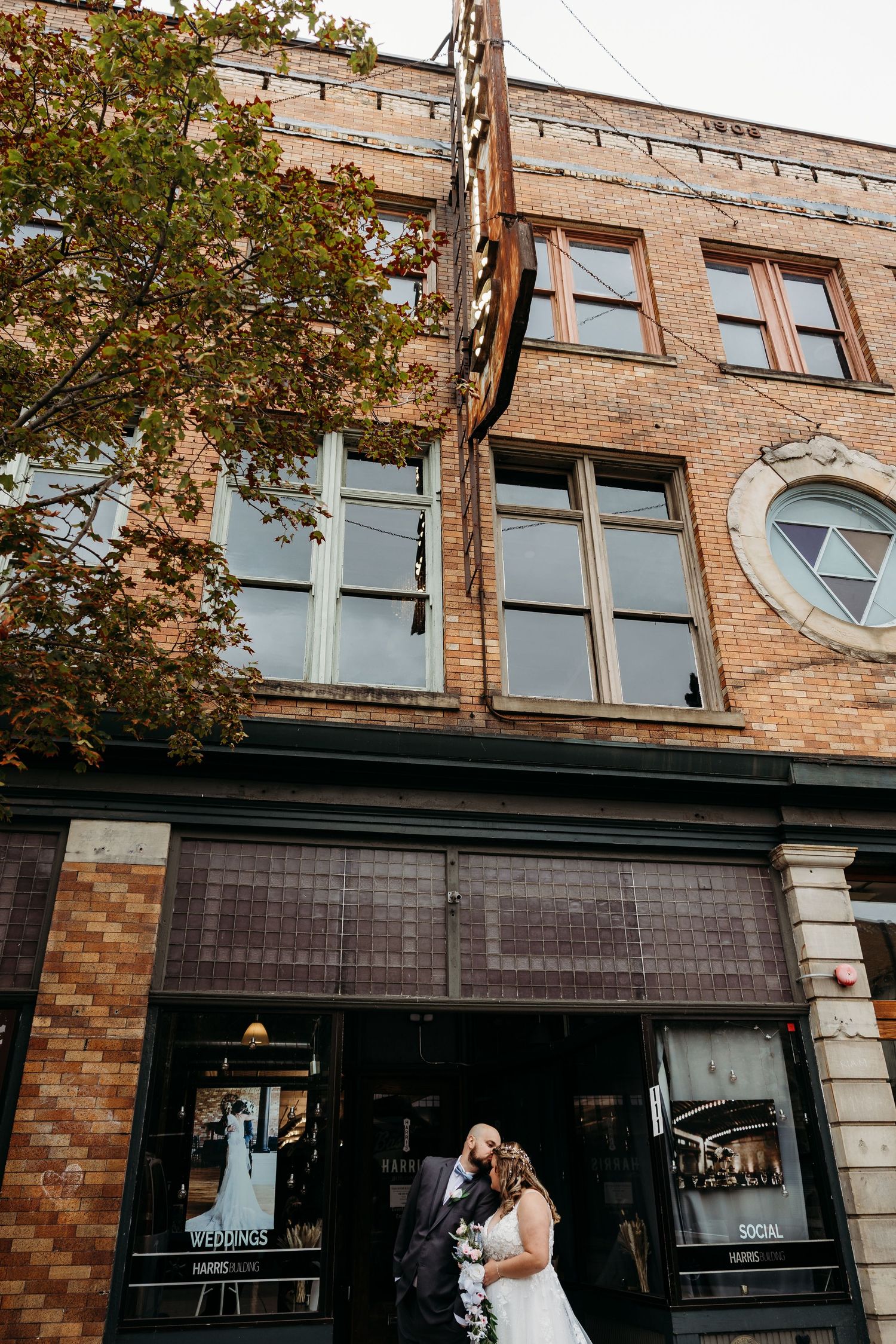 Bride and groom embrace outside a historic brick building with large windows on their wedding day.