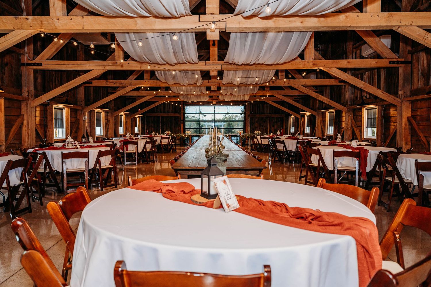 Rustic barn wedding reception with orange table runners, white linens, wooden chairs, and string lights under draped ceiling.