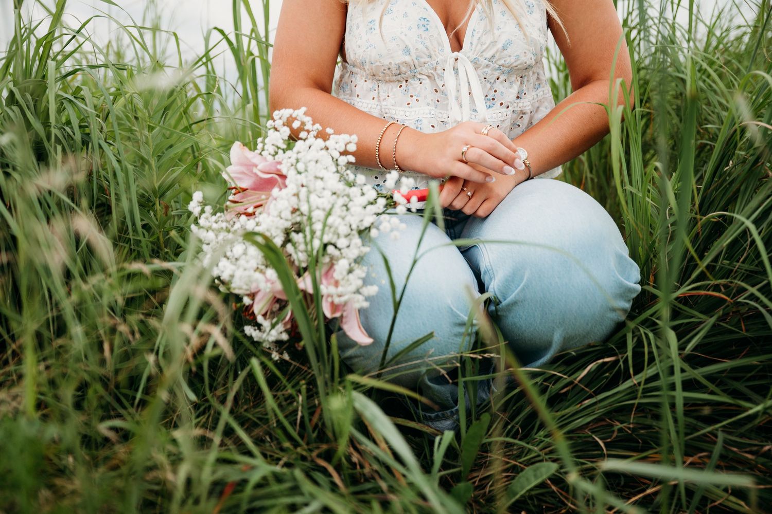 Person sitting in tall grass wearing white top and jeans holding flowers.
