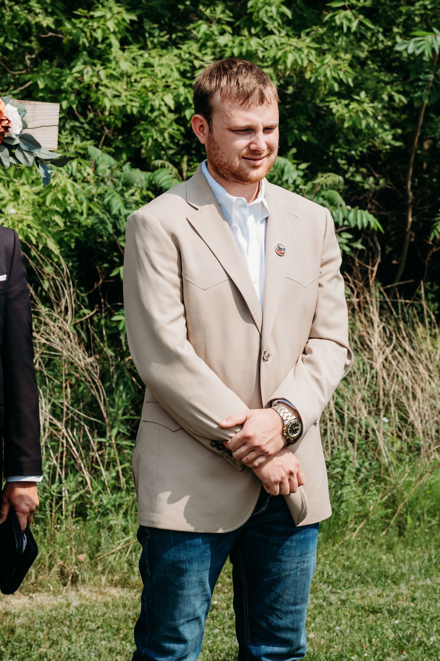 A man in a light beige blazer and blue jeans stands outdoors against a backdrop of green foliage and grass.