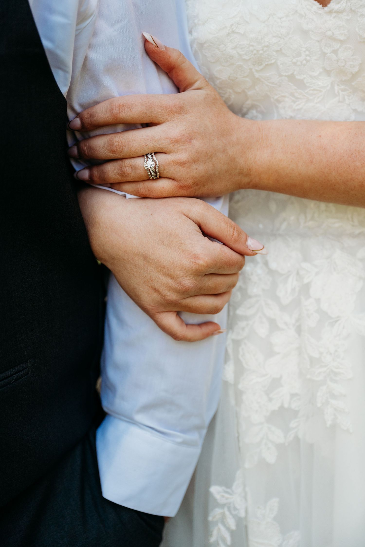 Close-up of hands with wedding rings embracing, showing white lace dress and light blue shirt sleeves.