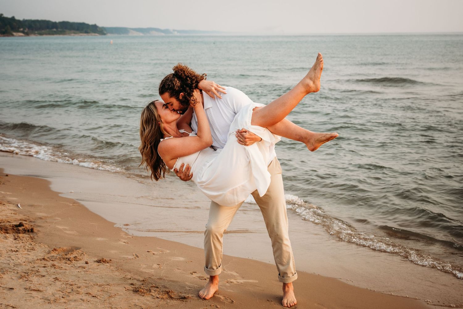 Romantic beach moment with couple in white clothing sharing a dip kiss at sunset.