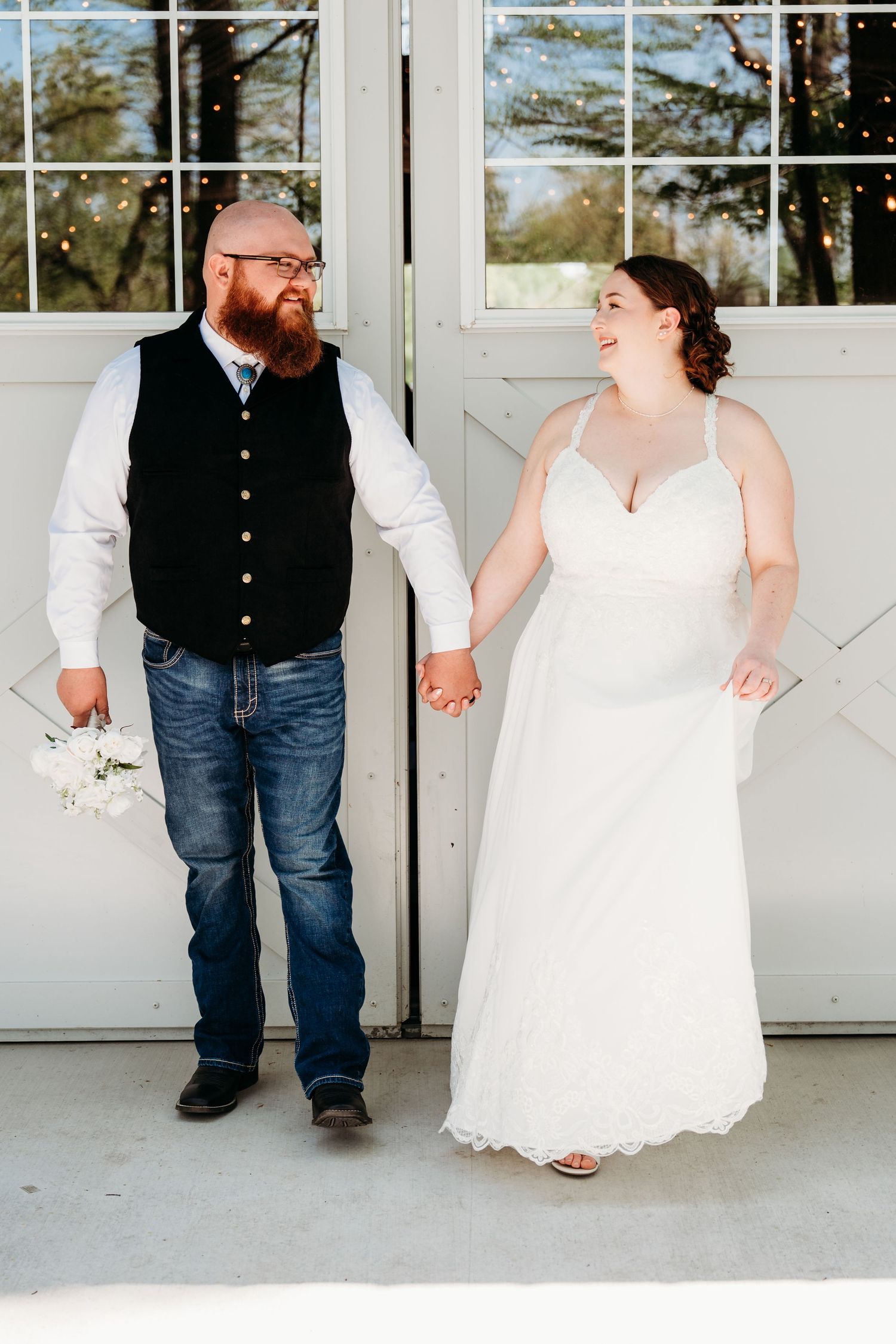 A couple in rustic wedding attire stands smiling together against white barn doors.