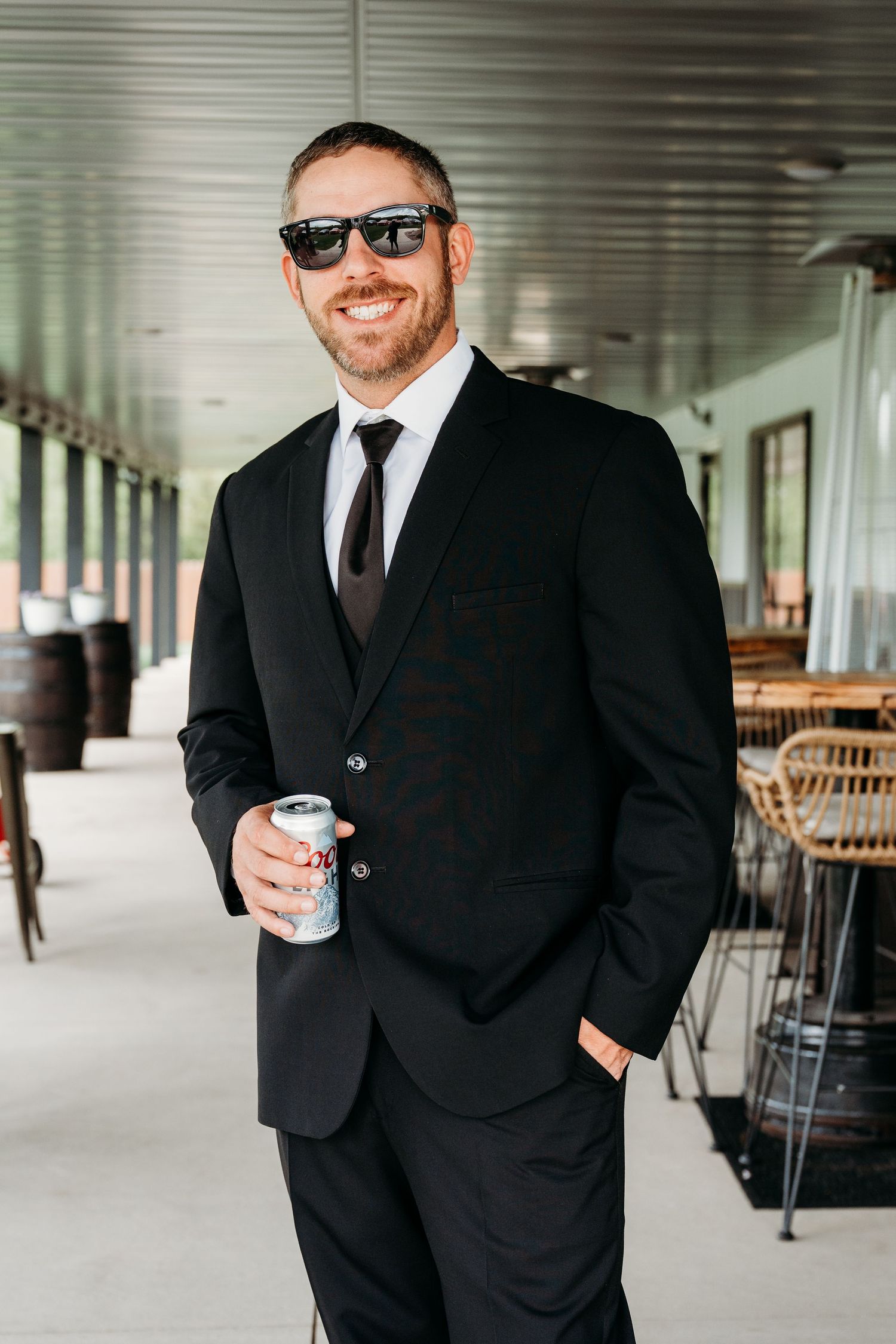 Groom in classic black suit and sunglasses stands on covered patio holding a drink.