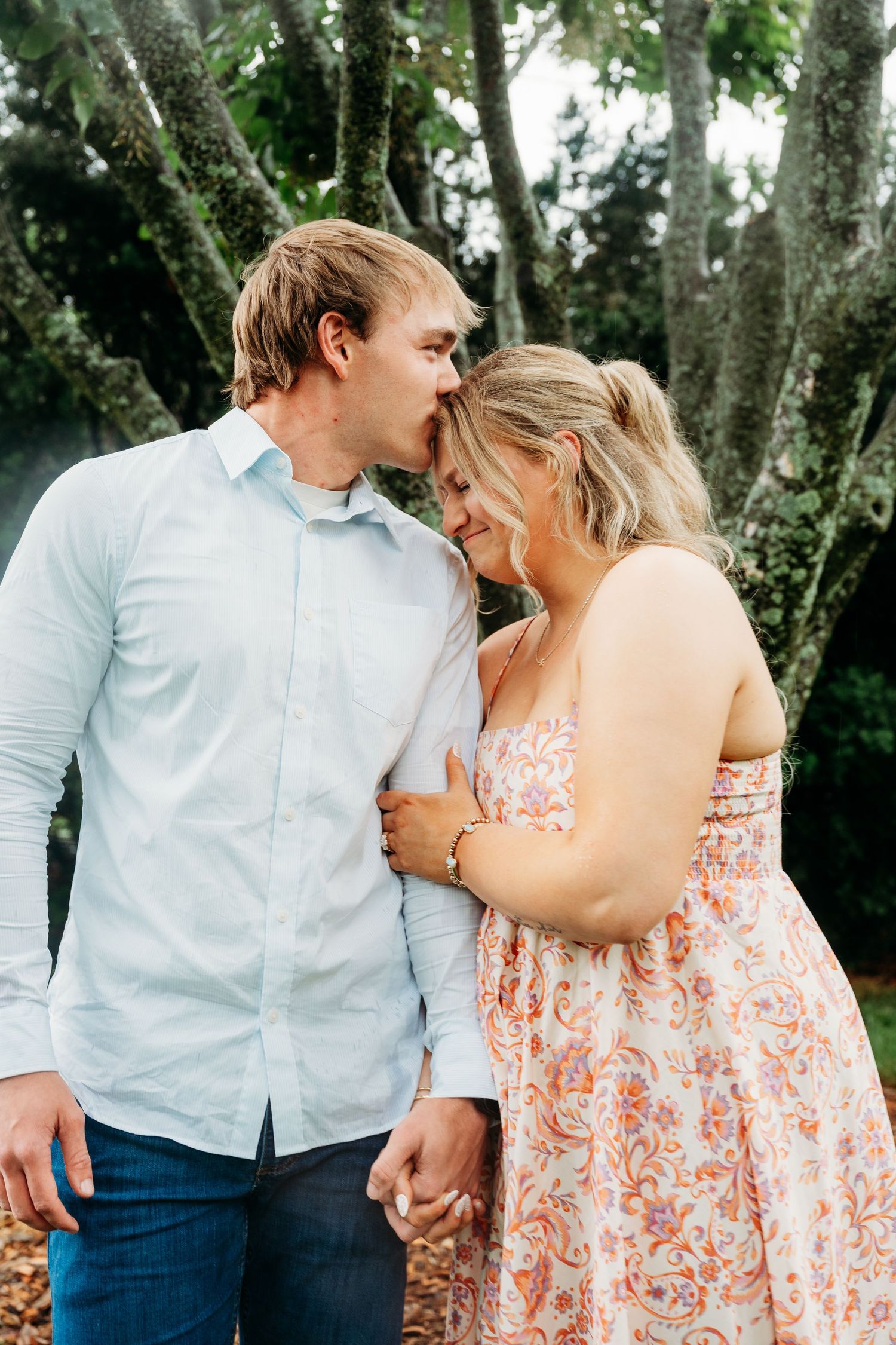 Couple sharing a tender moment outdoors, man kissing woman's forehead near mossy trees.