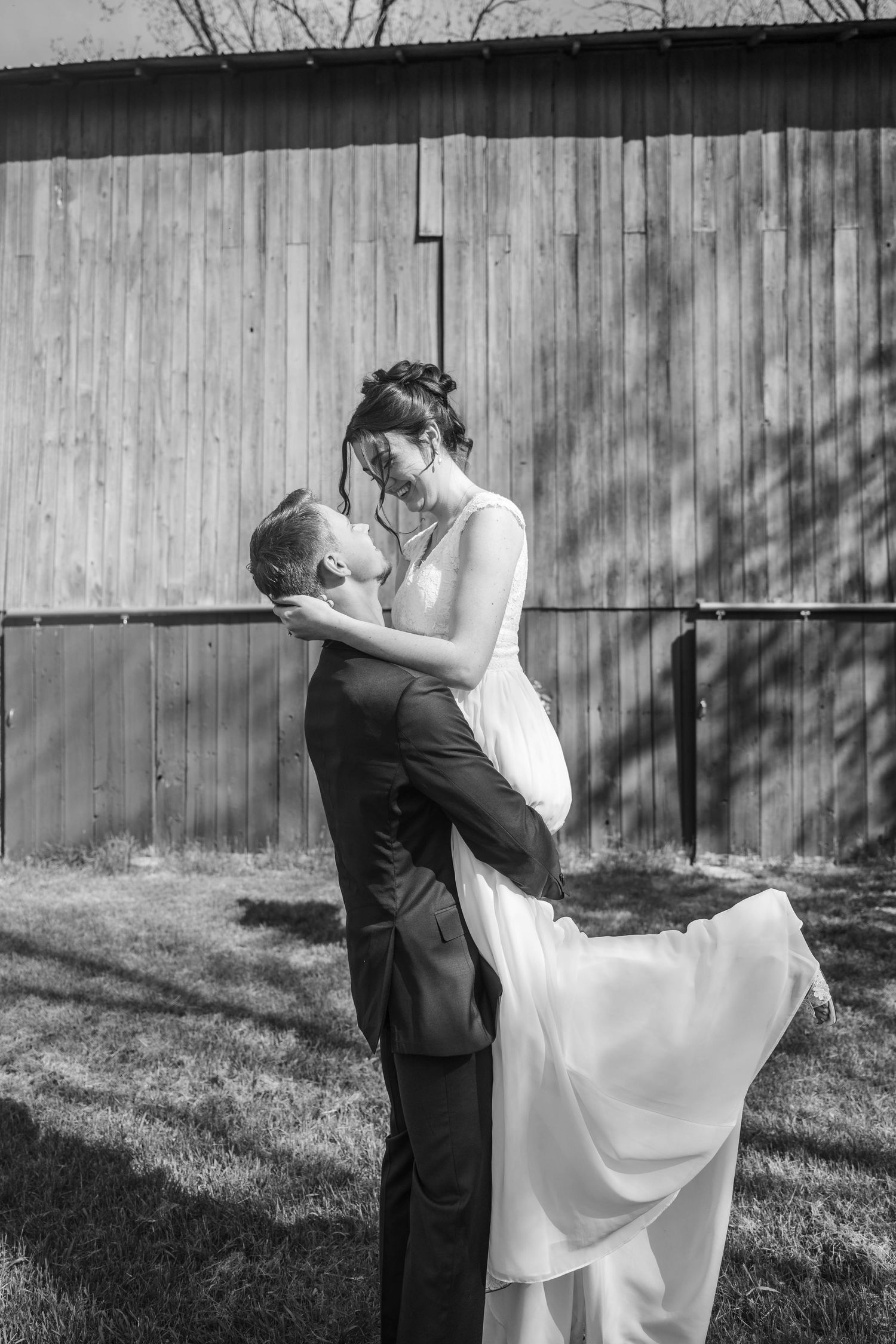Black and white photo of groom lifting bride in wedding dress outside rustic barn, sharing romantic moment.