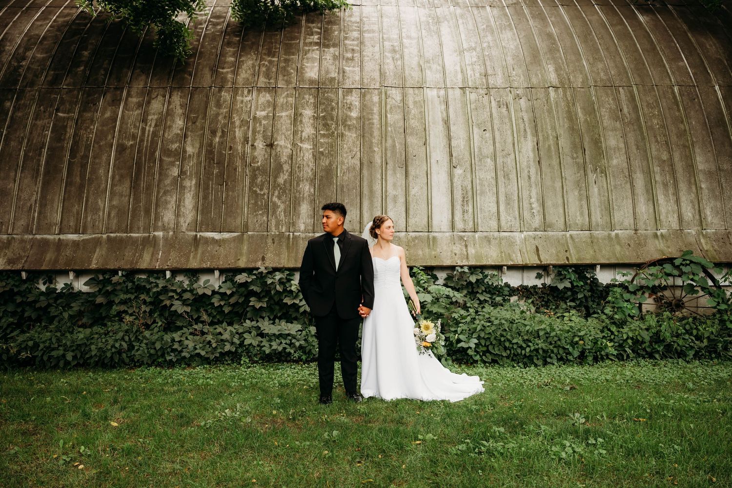 Bride and groom holding hands in front of a rustic wooden barn surrounded by lush greenery on their wedding day.