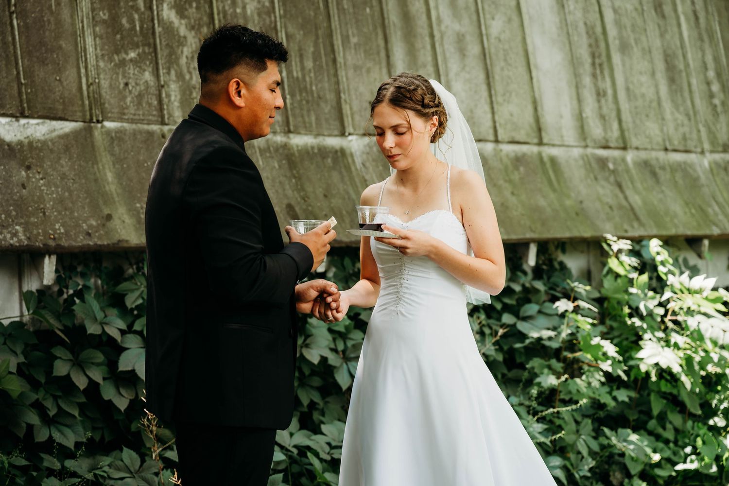 A bride in a white gown and veil exchanges rings with a groom in a black suit during an outdoor wedding ceremony.