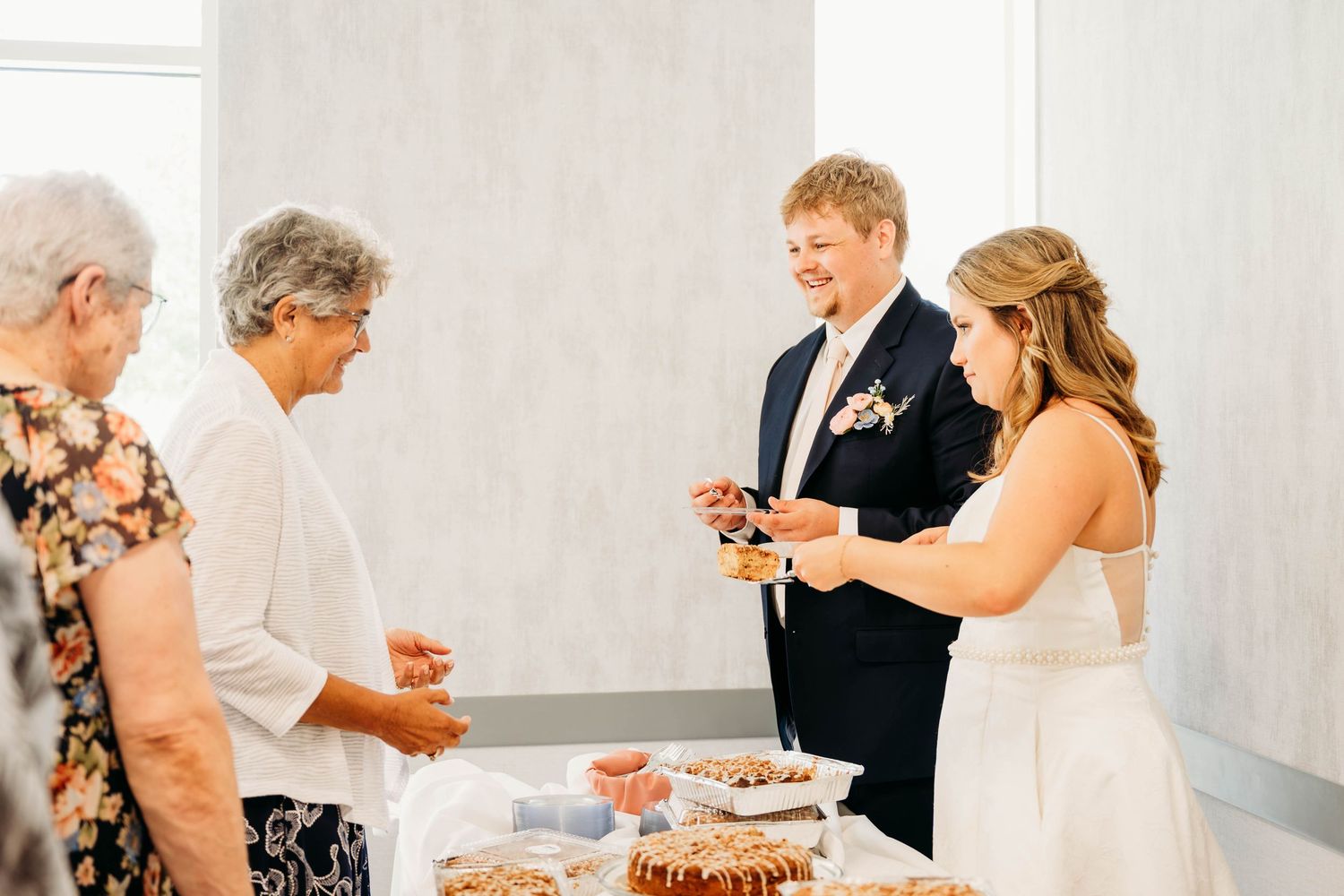 Bride and groom sharing wedding cake with elderly guests at a reception dessert table.