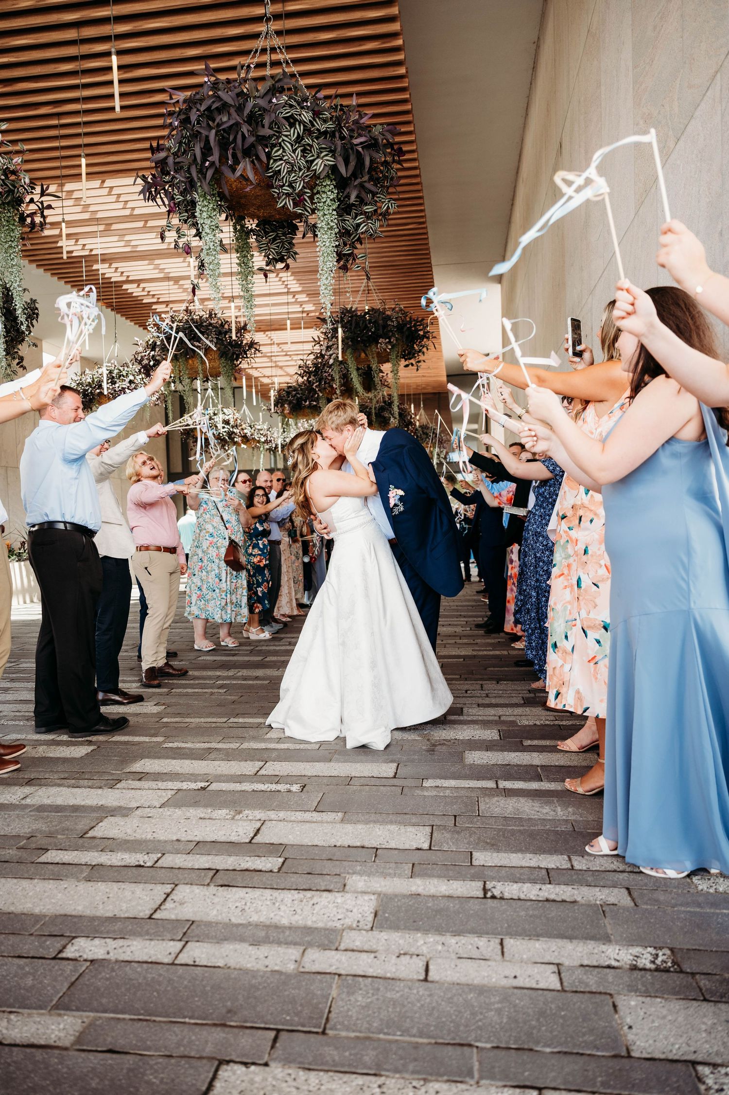 Bride and groom share a kiss during their wedding send-off, surrounded by guests holding ribbon wands outdoors.