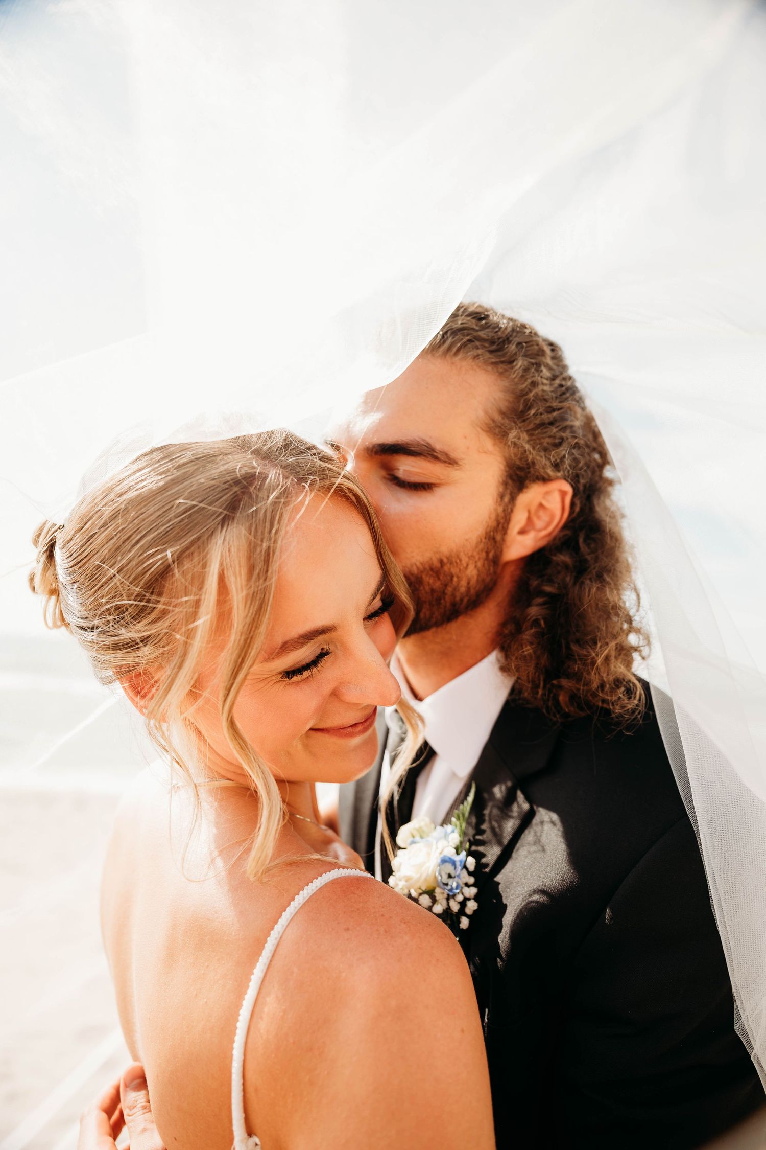 Groom kissing bride on cheek under veil at outdoor wedding, bride smiling in spaghetti strap gown.