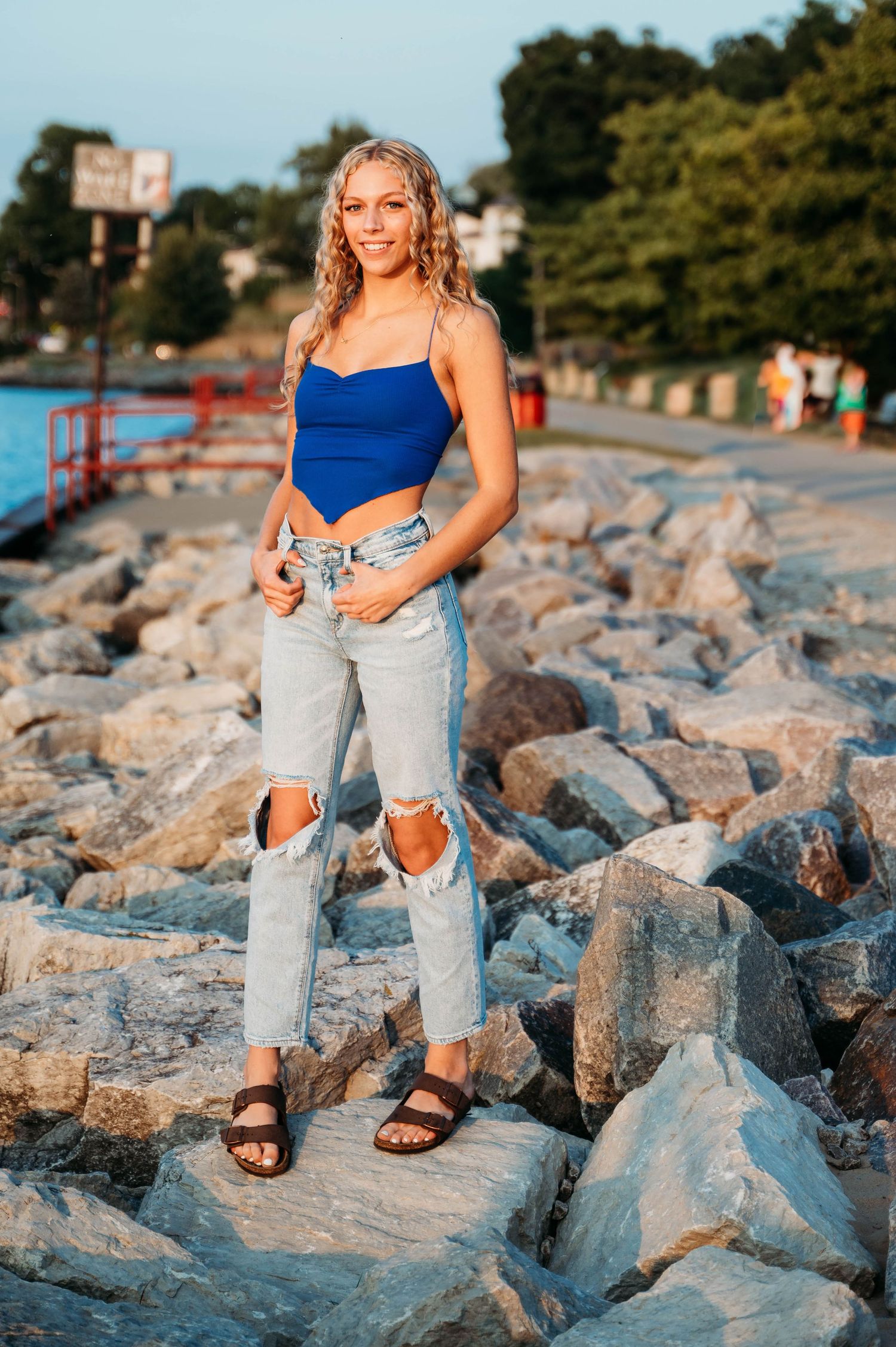 Person wearing blue bodysuit and ripped jeans poses on rocky shoreline at sunset with trees in background.