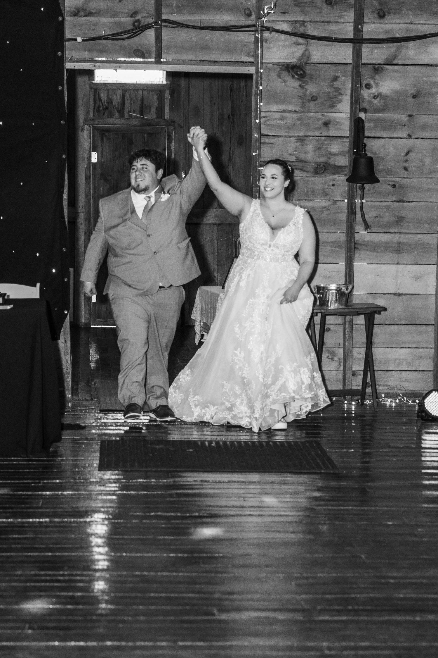 A couple makes their grand entrance at a rustic barn wedding venue, holding hands raised in celebration on a polished wood floor.