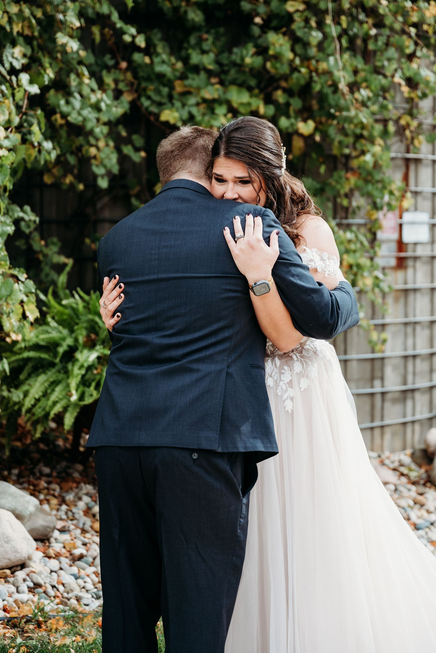 Bride in white lace gown embraces groom in navy suit during intimate outdoor garden wedding ceremony.