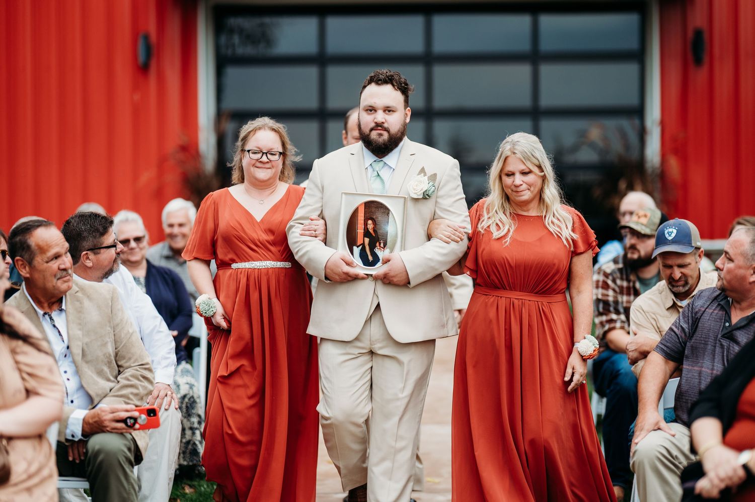 Wedding party in front of a bright red barn with guests in coral-colored attire.