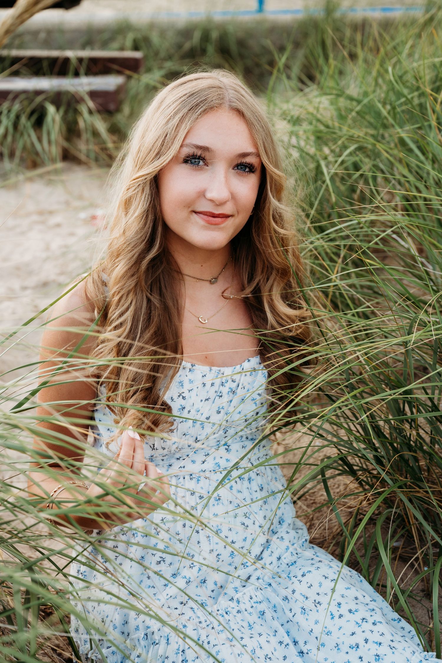 A smiling figure in a blue floral dress poses gracefully among tall beach grass during an outdoor photo session.