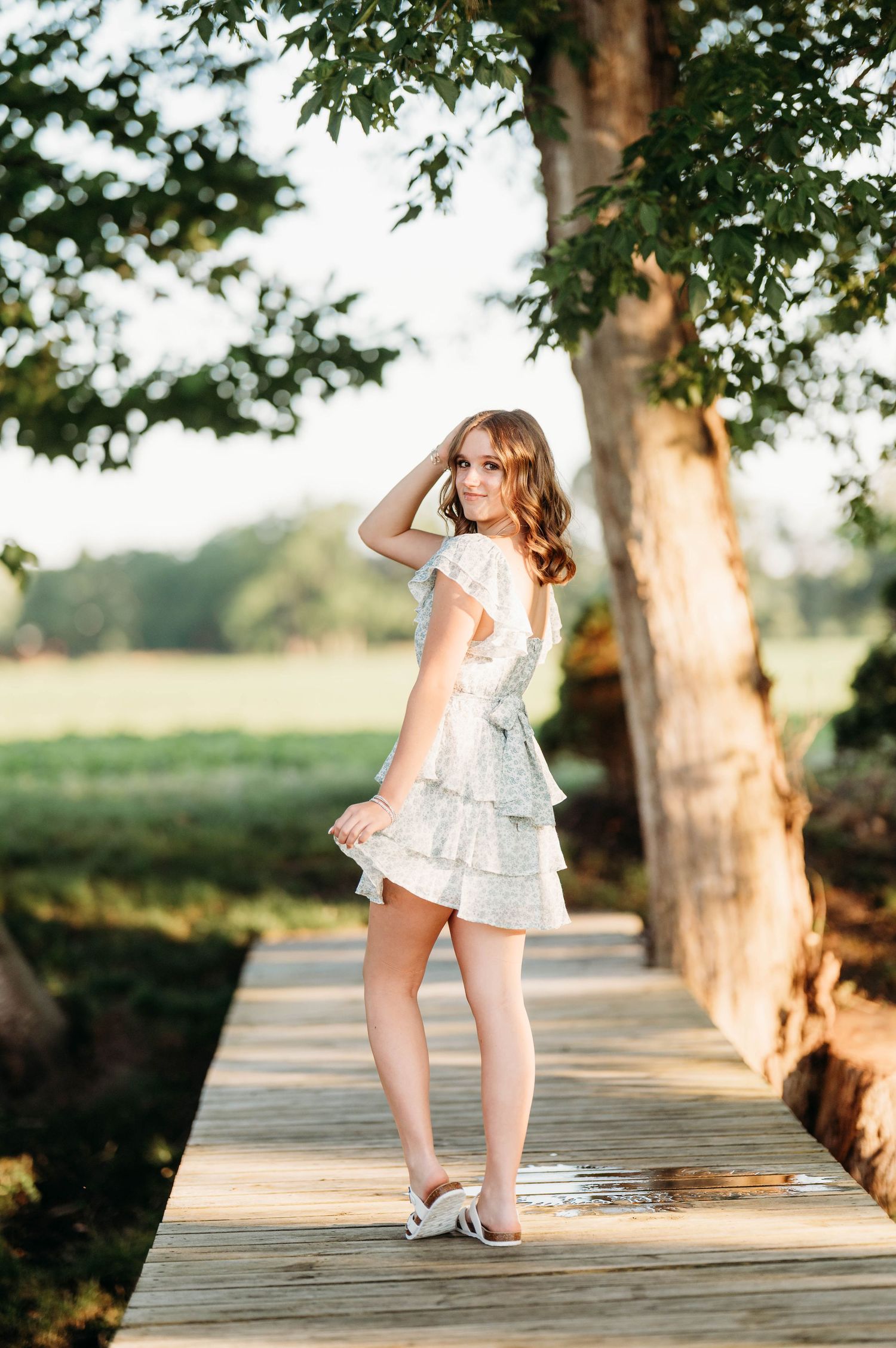 Young woman in white tiered lace dress poses on wooden boardwalk surrounded by lush green trees in golden sunlight.