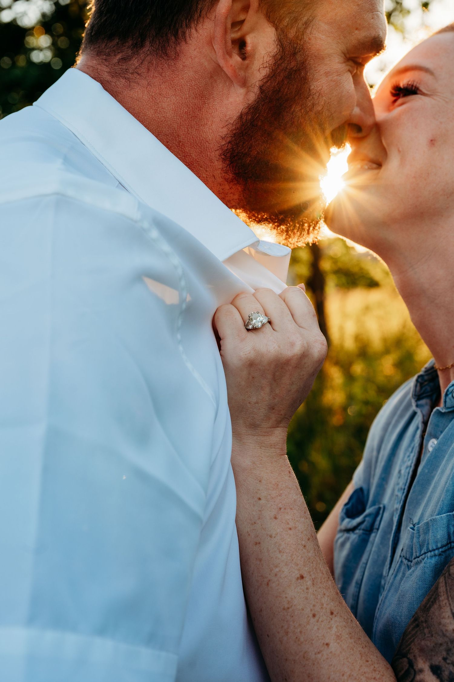 Couple sharing a romantic sunset kiss, woman wearing diamond engagement ring, golden backlight glowing between them outdoors.