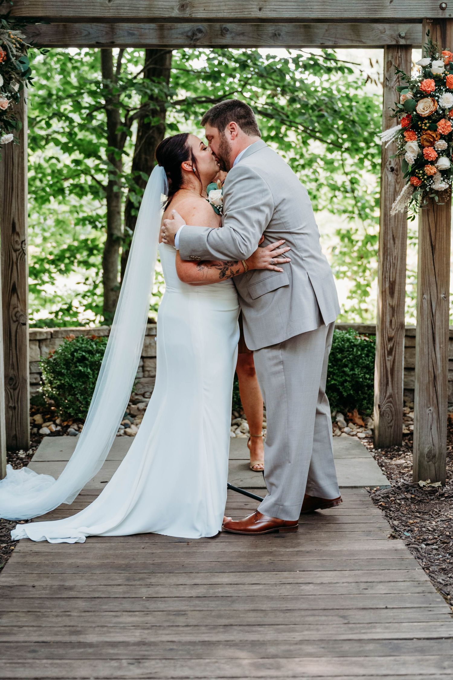 A couple shares a romantic kiss during their outdoor wedding ceremony under a wooden pergola decorated with flowers.