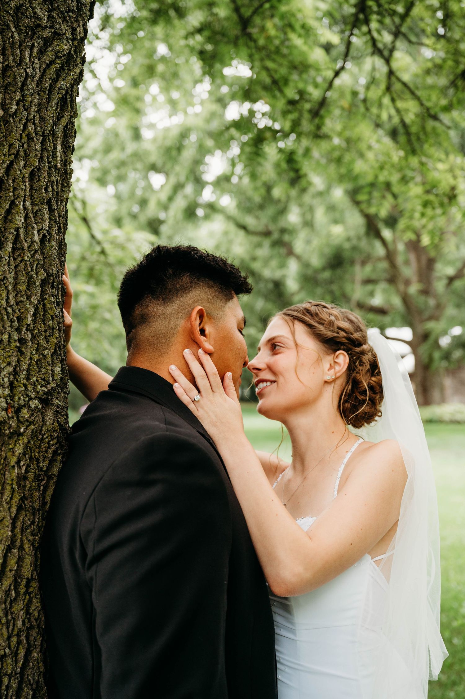 Bride and groom share a romantic moment by a tree, with bride in white gown and veil smiling at her partner.