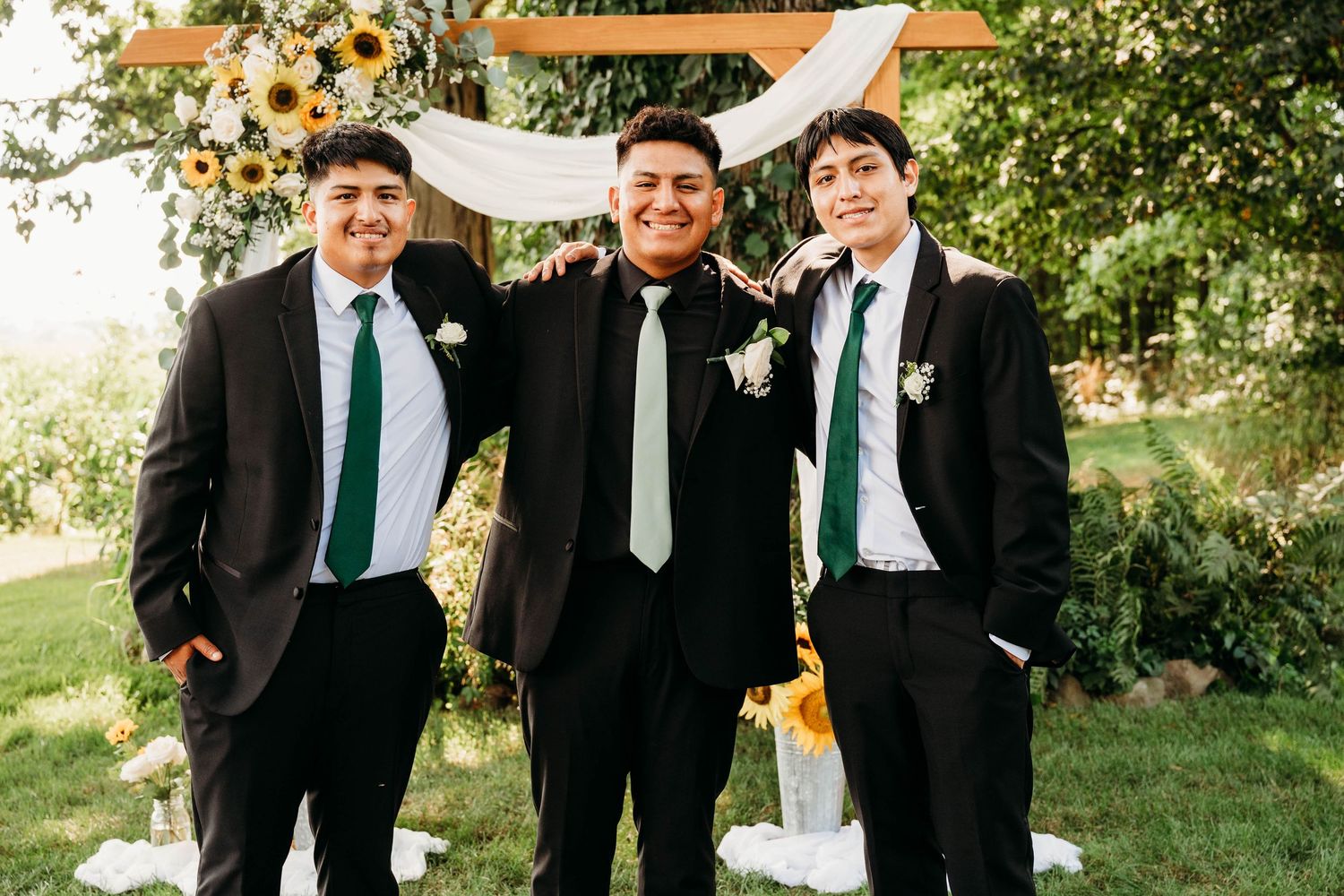 Three groomsmen in black suits with green ties pose together at an outdoor wedding ceremony.
