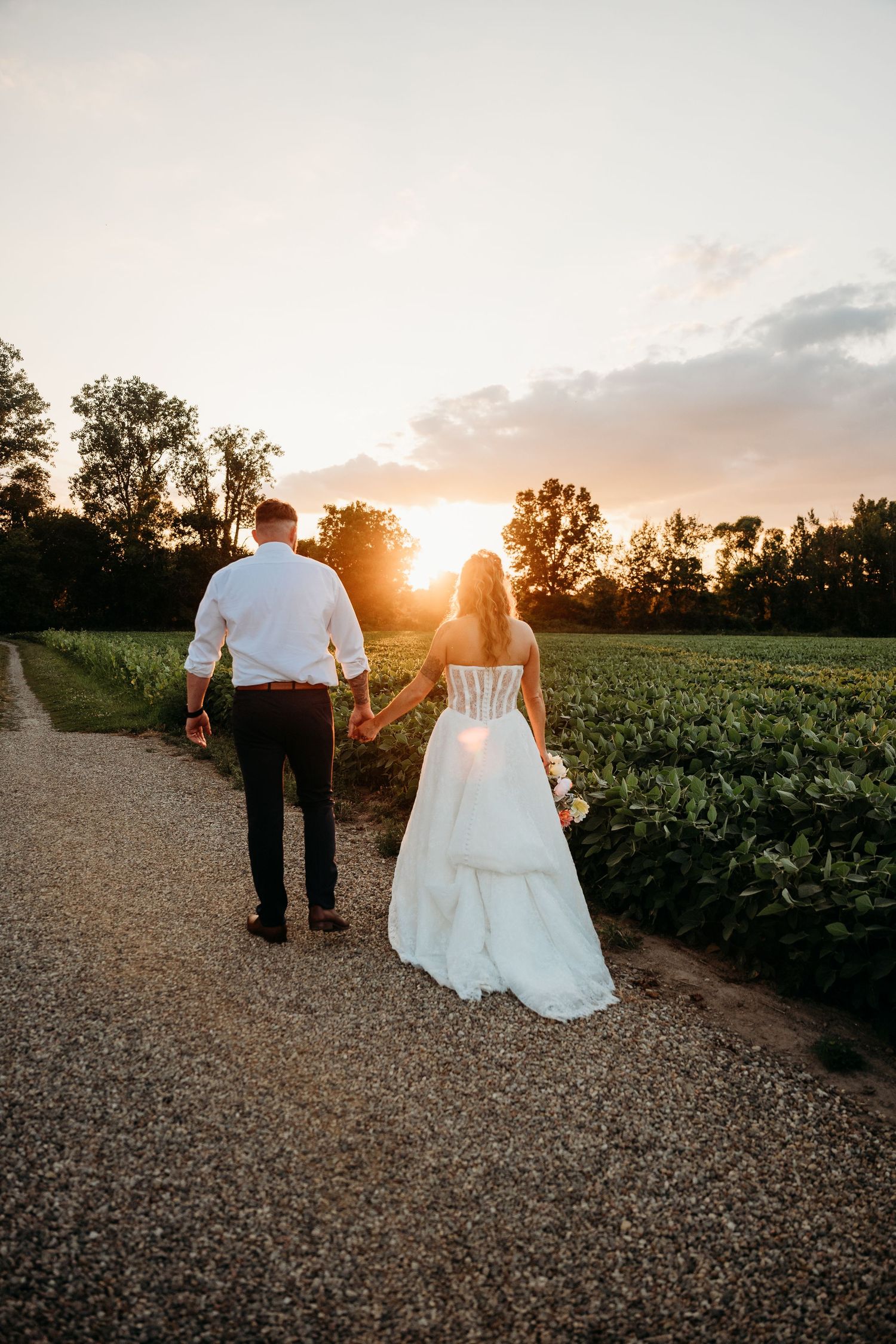 Bride and groom walk hand-in-hand down a gravel path at sunset, surrounded by lush green fields.