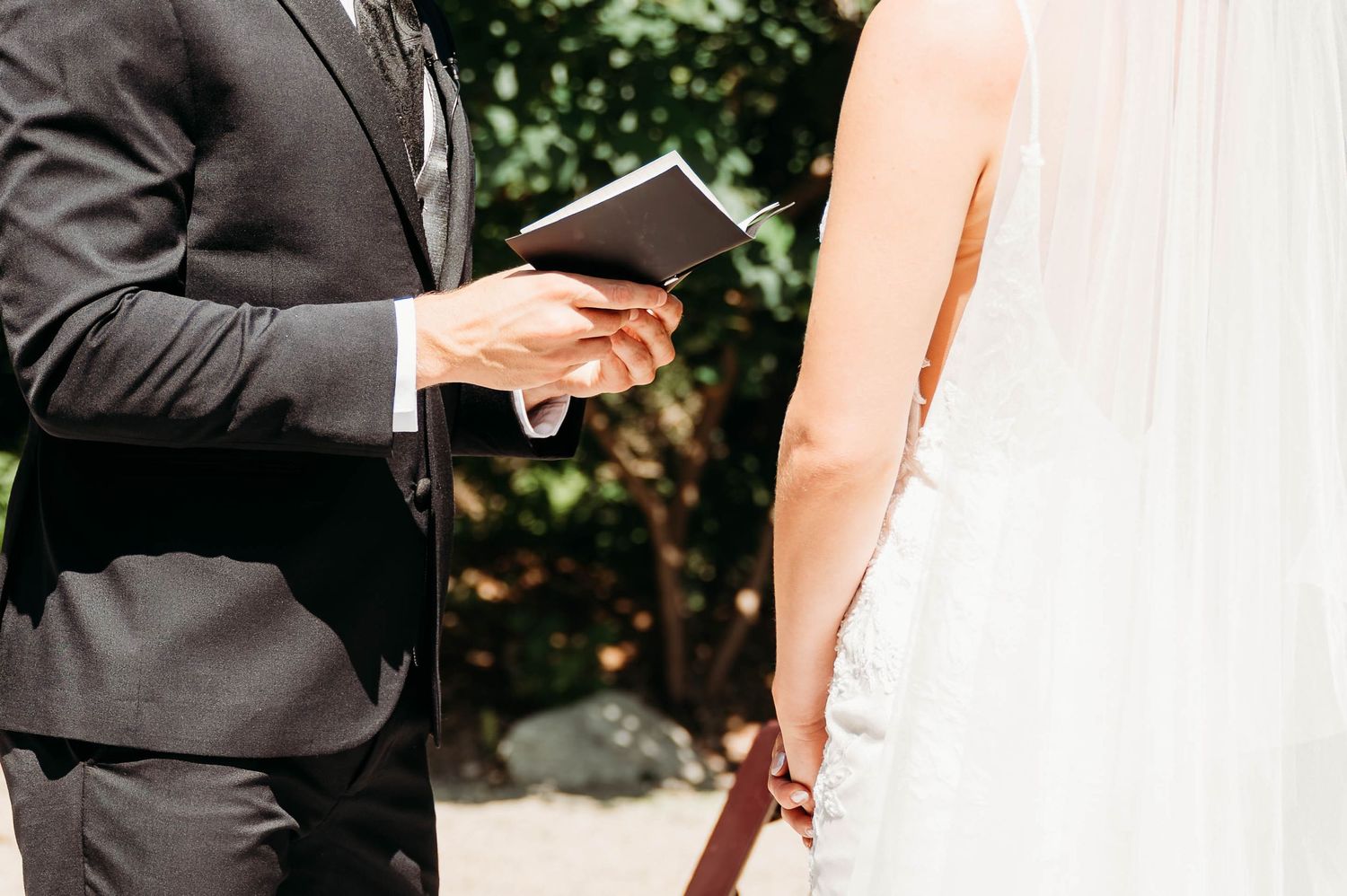 Groom in black suit reads vows from booklet to bride in white lace wedding dress during outdoor ceremony.