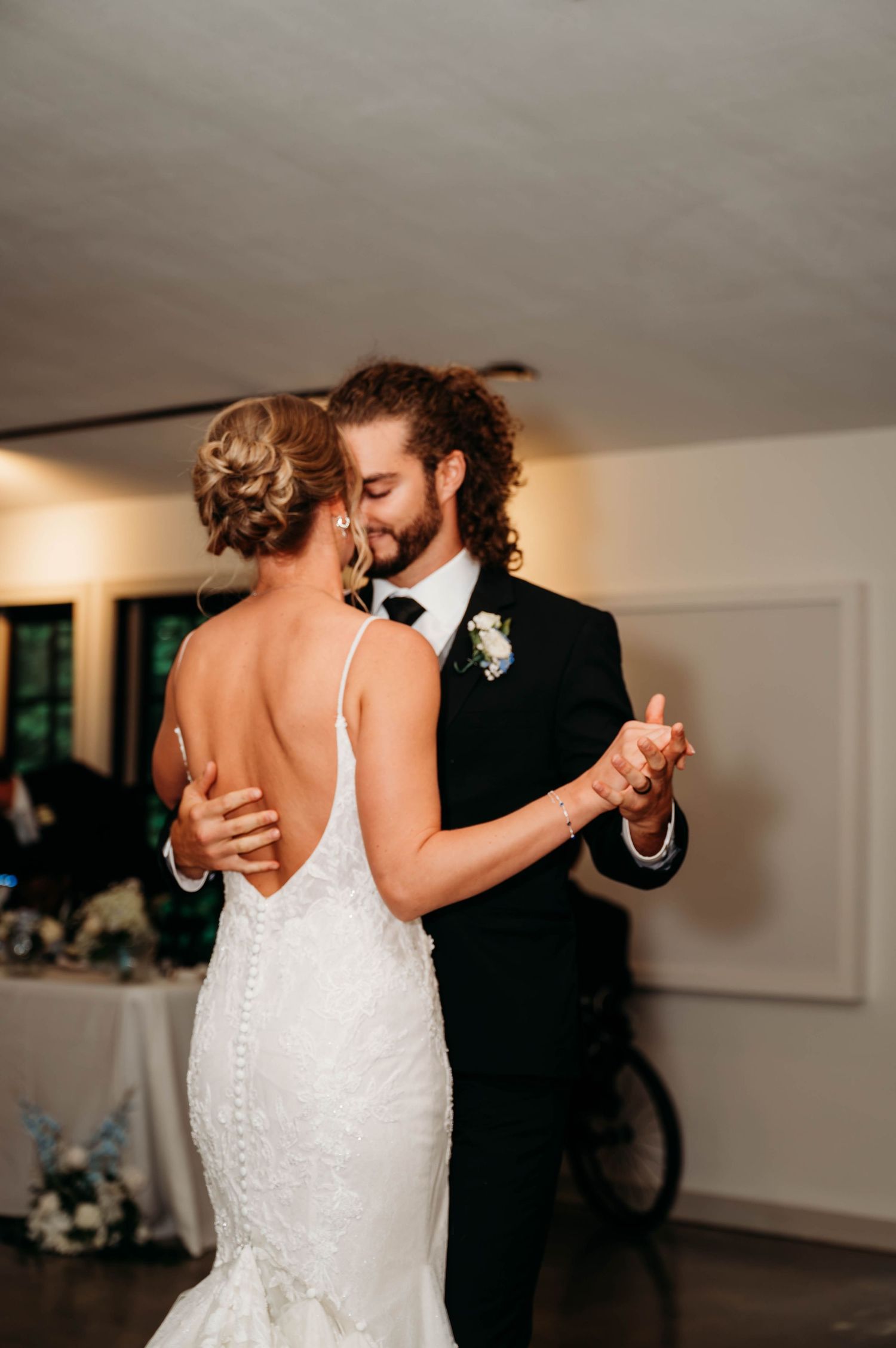 Bride and groom share their first dance, she in a backless lace gown, he in a black tuxedo with boutonniere.