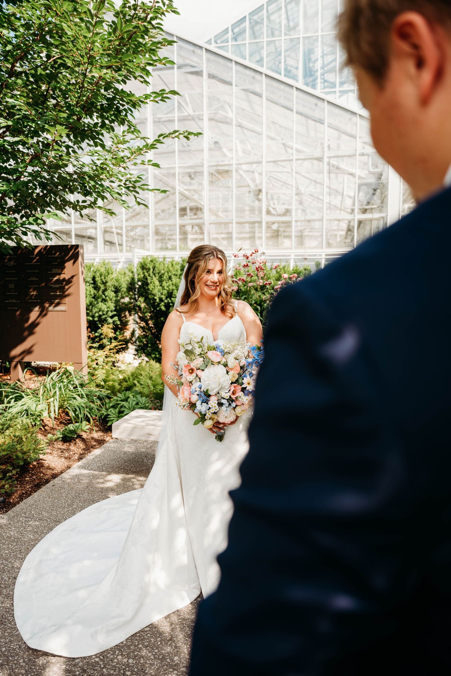 Bride in white gown holding colorful bouquet smiles at groom in navy suit outside greenhouse venue.