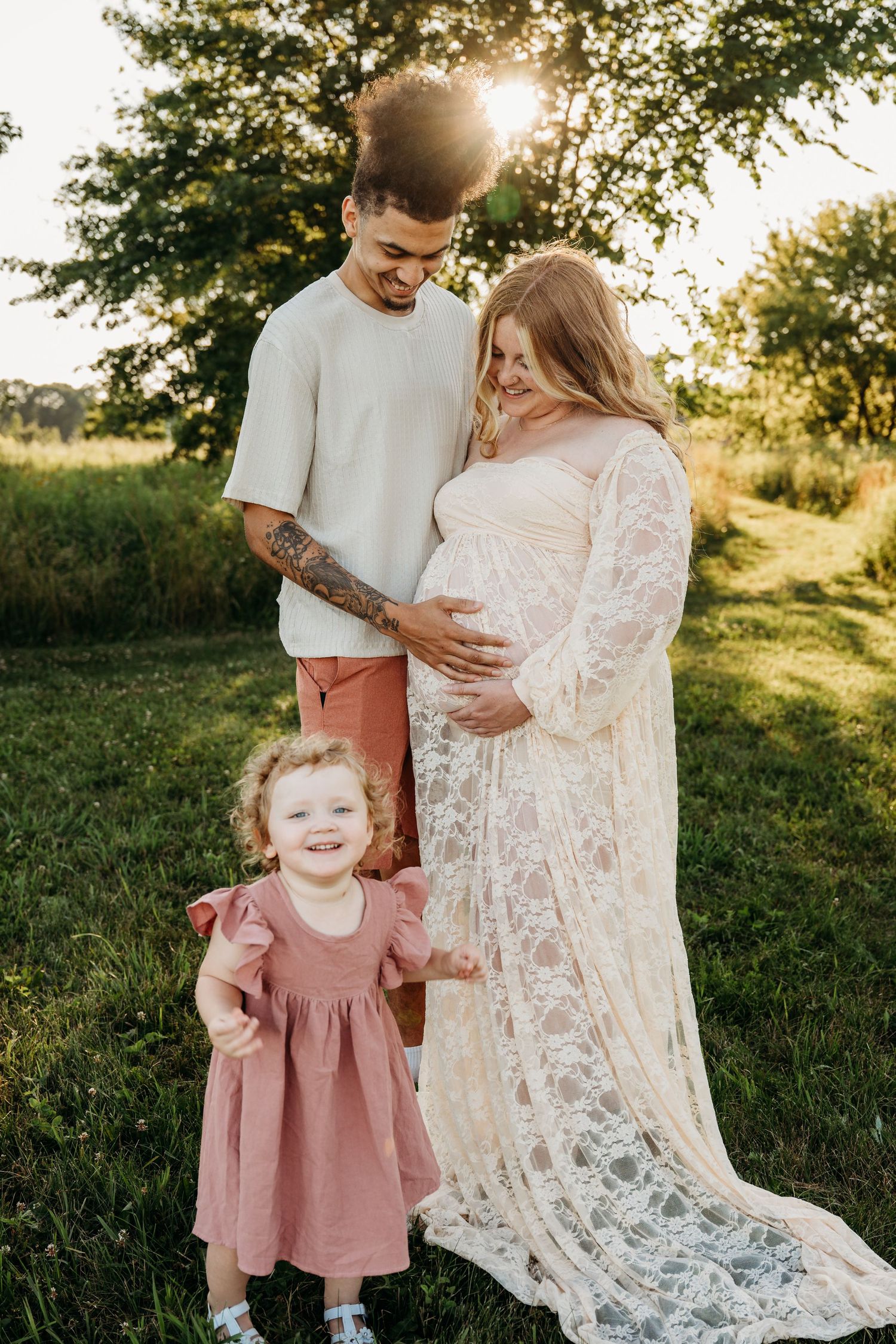 Family maternity photo in golden sunlight showing happy couple with toddler in matching mauve and cream outfits.