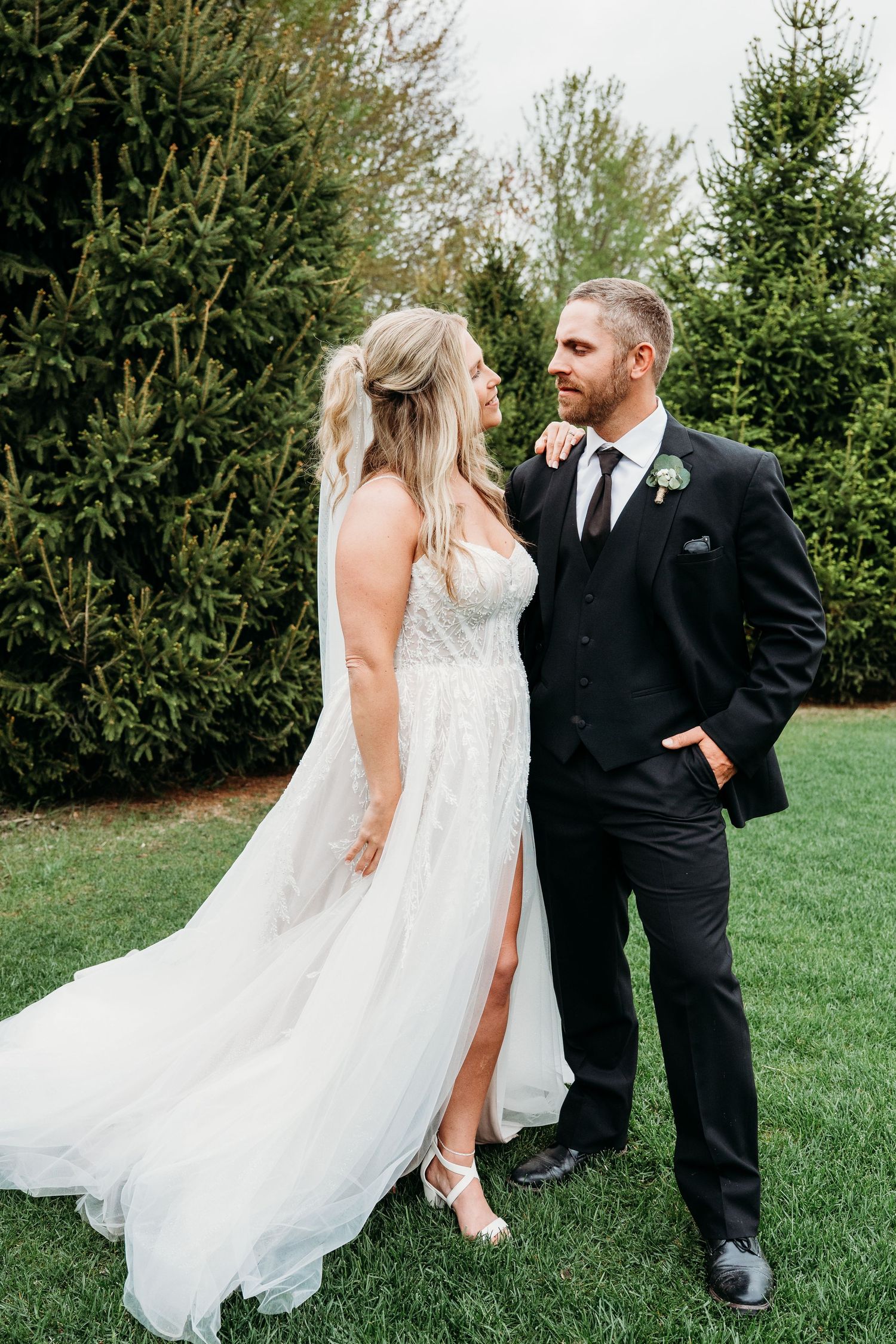 Elegant bride in lace gown with flowing veil gazes at groom in black suit amid evergreen trees outdoors.