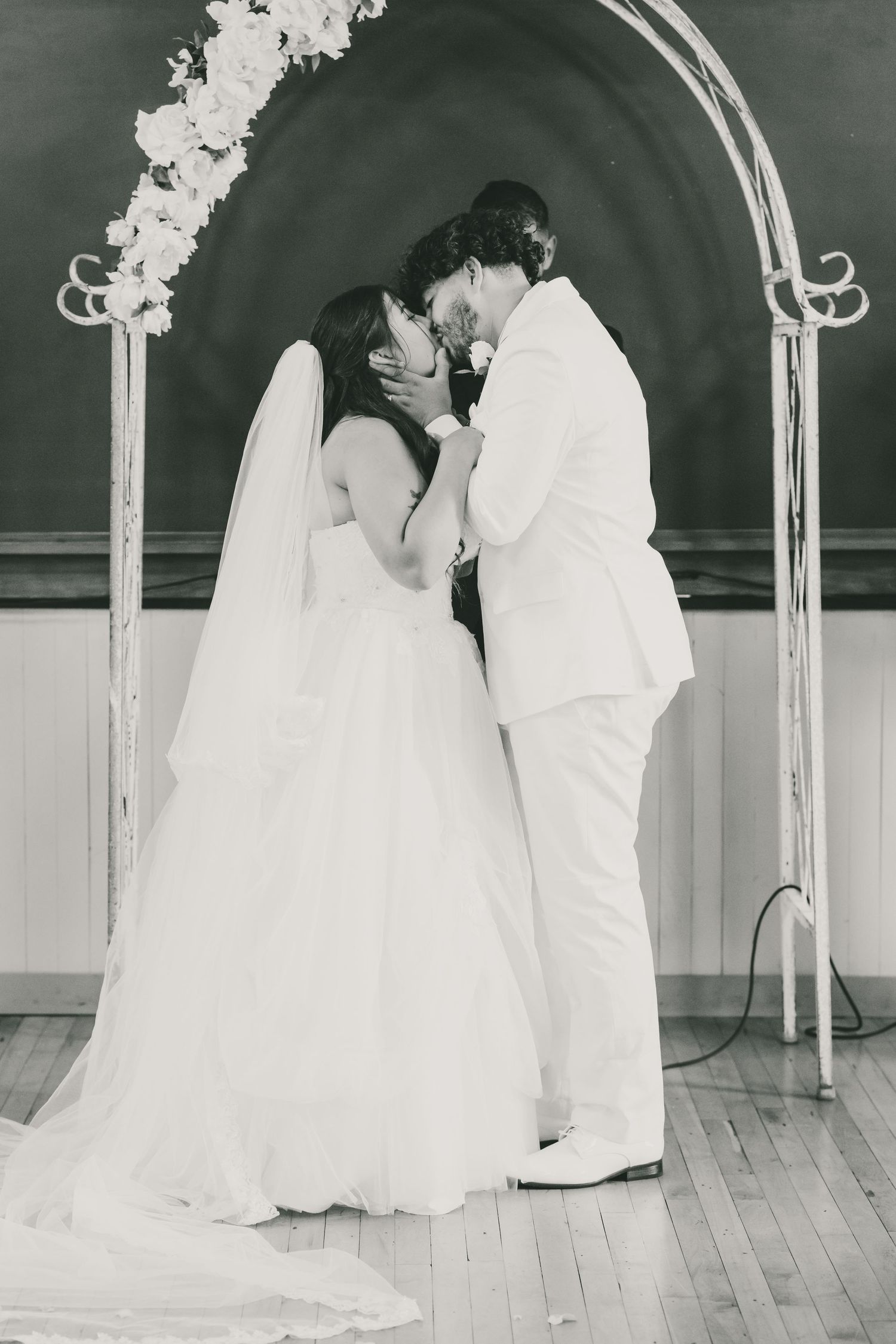 Black and white photo of bride and groom sharing first kiss under floral arch at indoor wedding ceremony. The Bell Flower Kalamazoo
