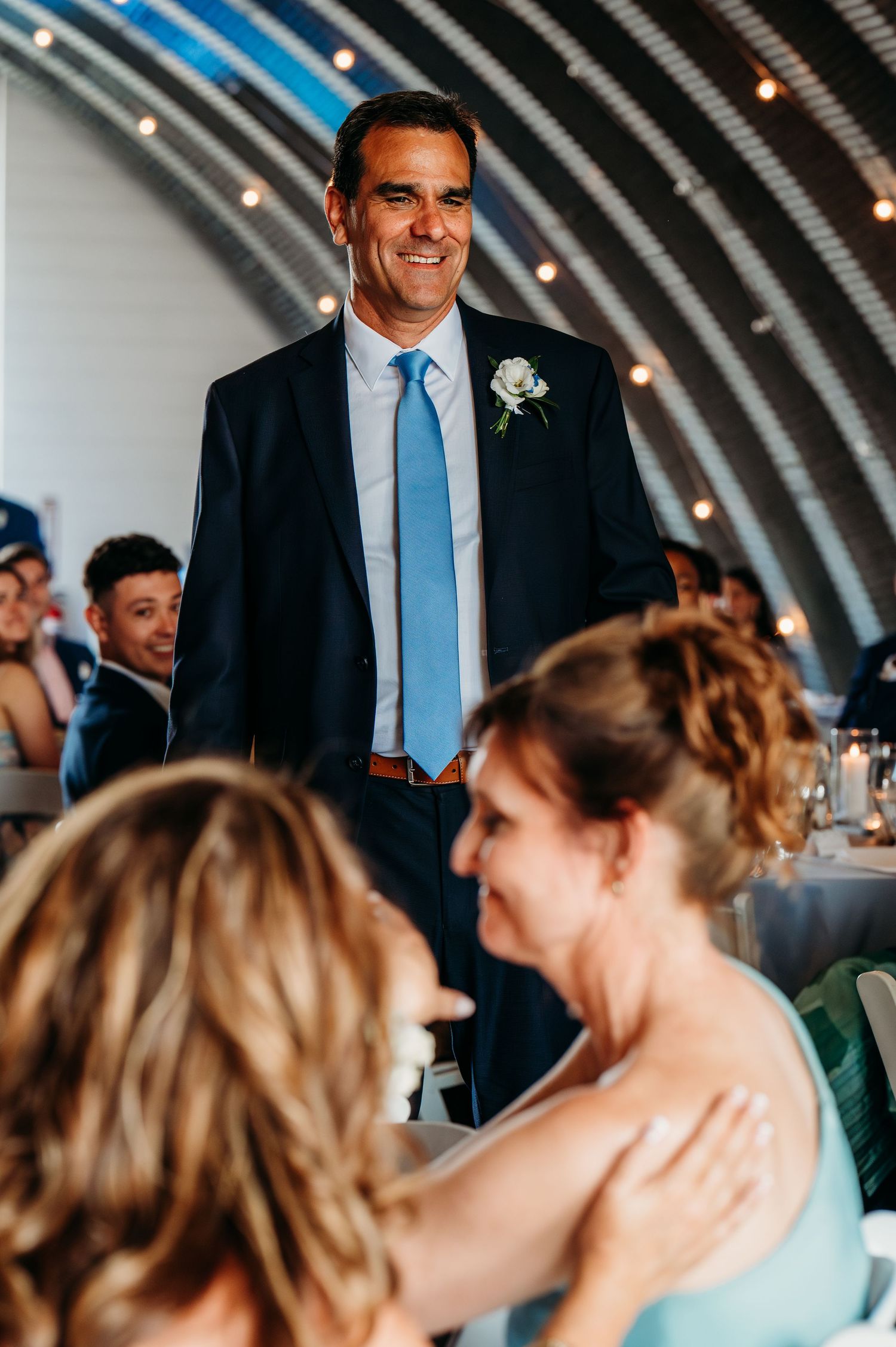 Man in navy suit and blue tie smiling at wedding reception with guests seated at tables in elegant venue.