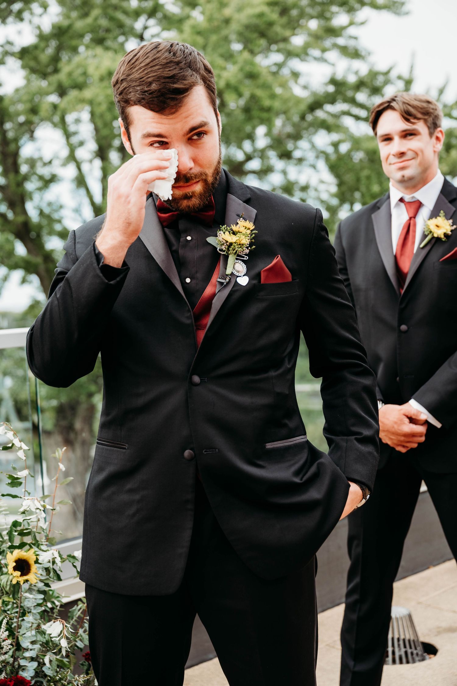 Emotional groom wiping tears at wedding altar in black tuxedo with red bow tie and sunflower boutonniere.