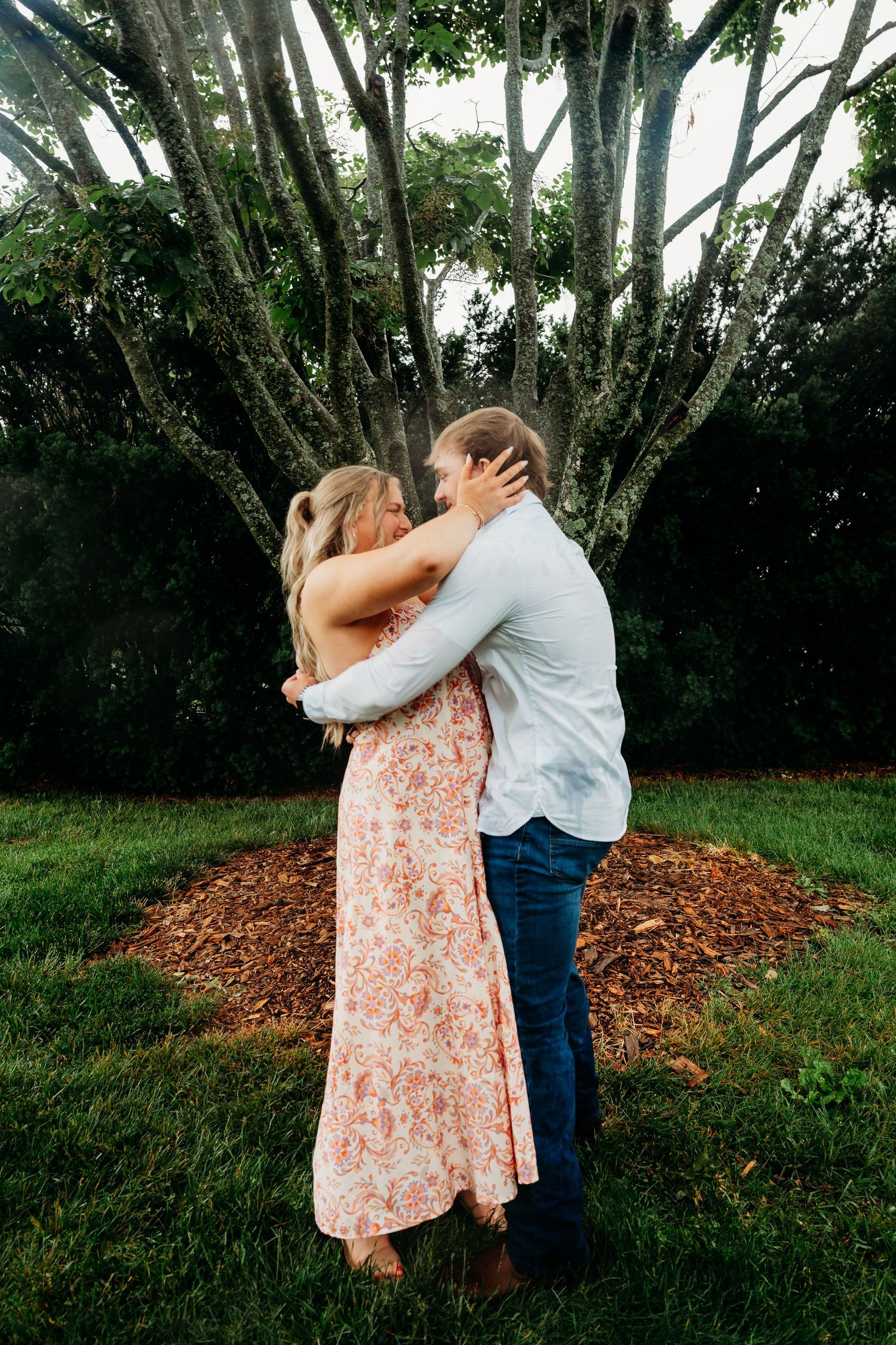 Couple embracing and kissing outdoors near a large tree, woman in floral dress and man in blue jeans.