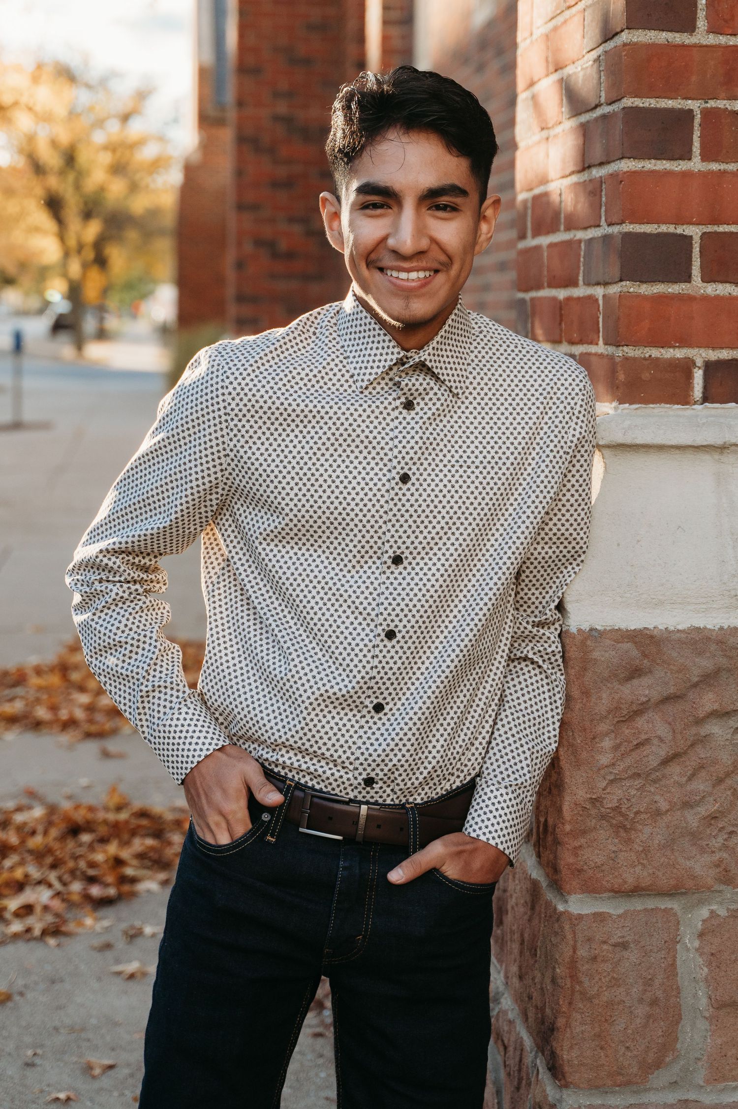 A person wearing a polka dot button-up shirt and dark jeans poses casually against a brick wall on an autumn day.