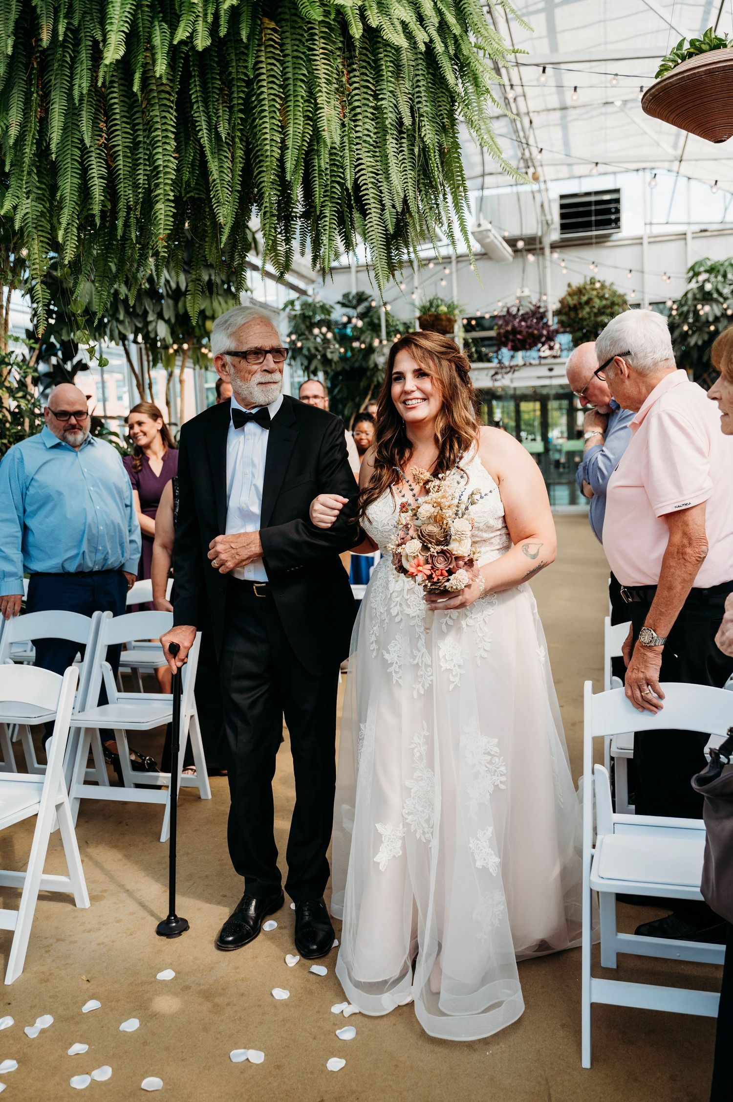 A wedding ceremony takes place in a greenhouse setting with hanging ferns and white folding chairs.