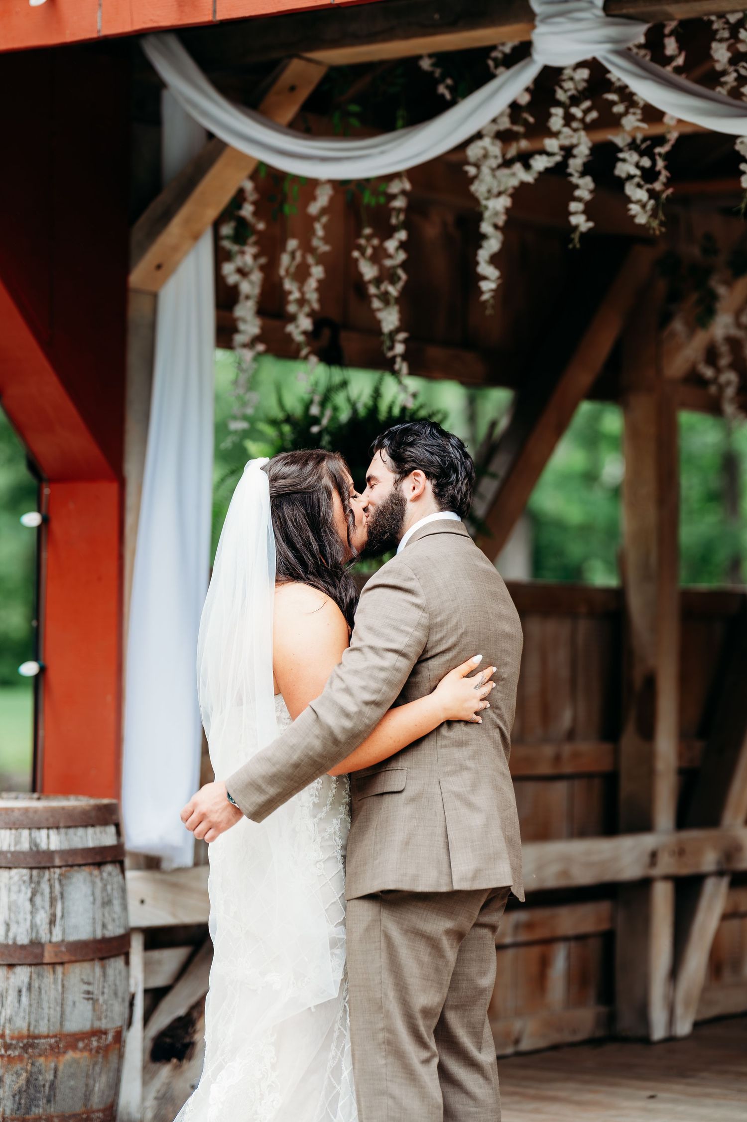 Bride and groom share first kiss under rustic wooden pavilion decorated with floral garlands at outdoor wedding.