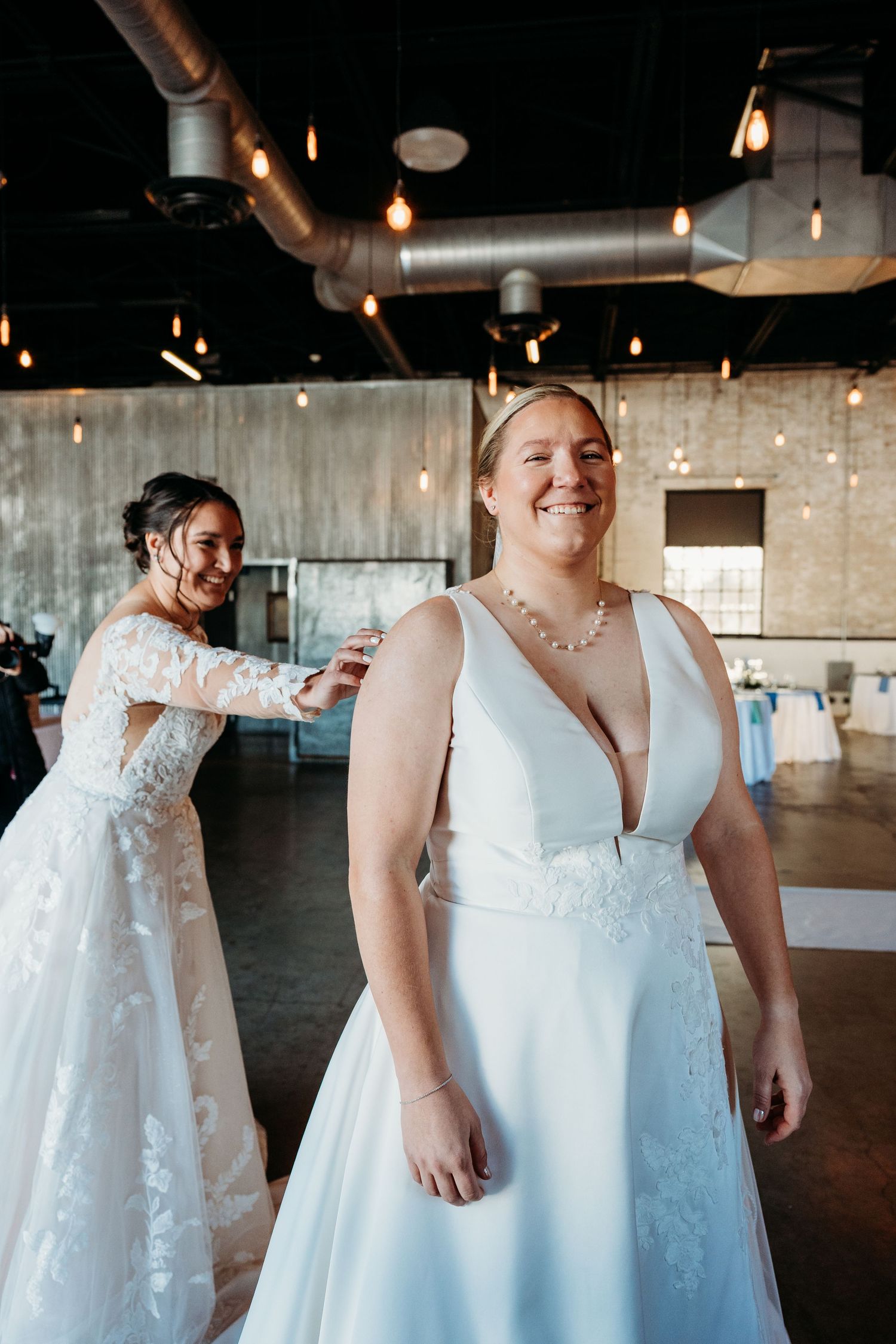 Two people wearing elegant white wedding dresses laugh together in an industrial-style venue with exposed ductwork and lights.
