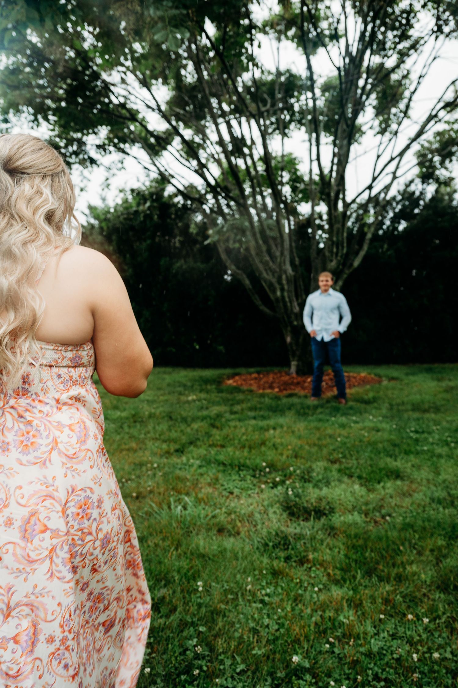 A woman in a floral dress walks toward a man in a blue shirt standing near a tree in a lush green yard.